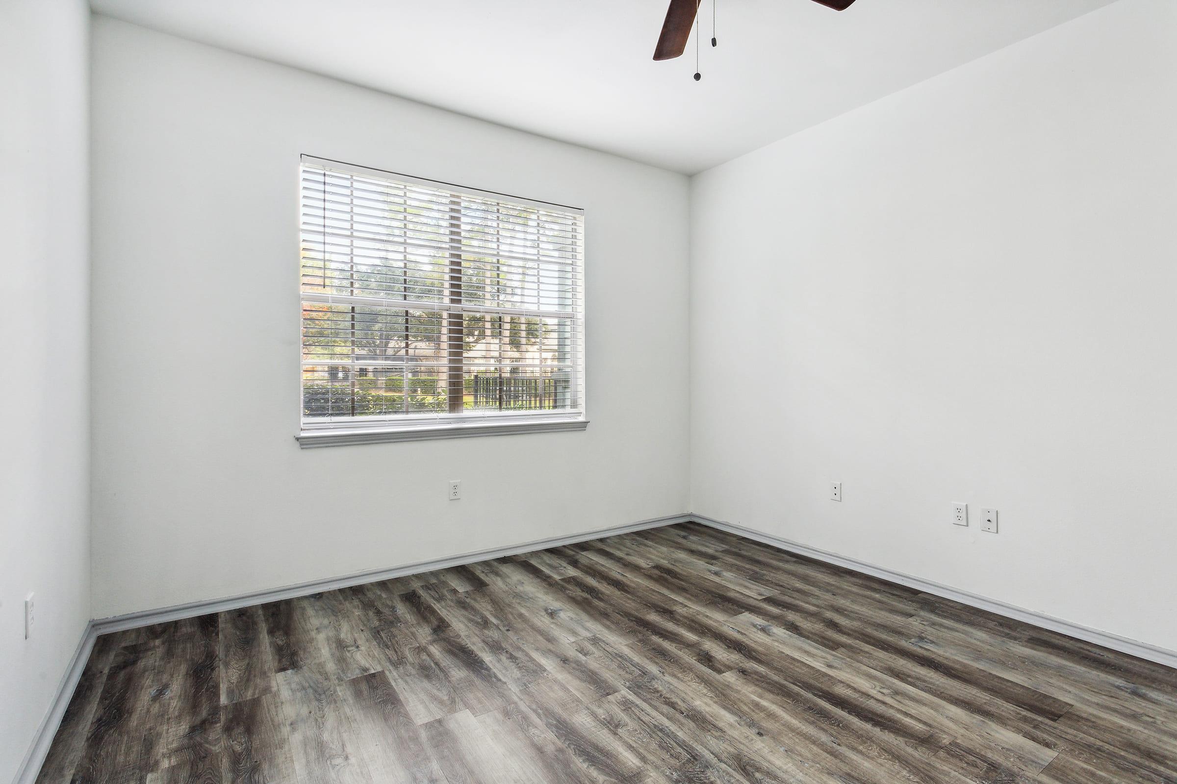 A bright, empty room with light-colored walls, a window featuring white blinds, and a ceiling fan. The floor is covered with wood-like laminate, and there is a small amount of natural light coming through the window, casting shadows on the floor. The space is uncluttered and ready for furnishing.