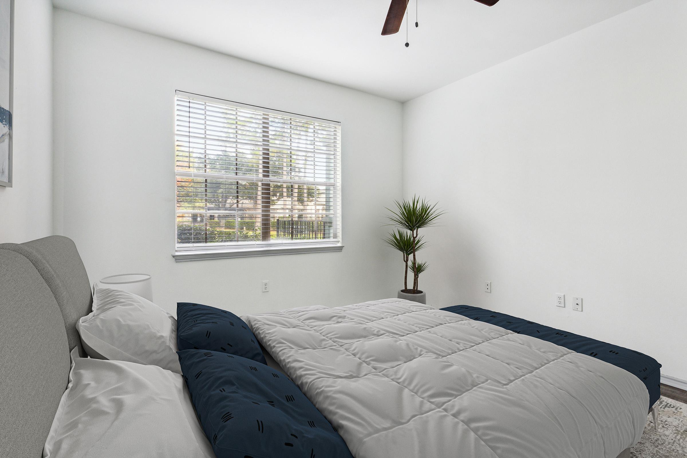A well-lit bedroom featuring a bed with a blue and white comforter, a nightstand with a lamp, and a small plant by the window. The walls are painted white, and there are light curtains allowing natural light to filter in from the outside.