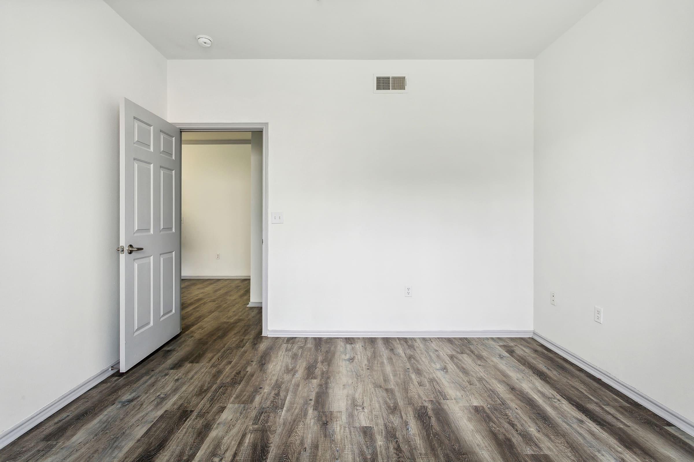 A spacious, empty room featuring light-colored walls and a wooden floor. A closed white door is visible on the left, while an open door leads to another room at the back. Natural light brightens the space, highlighting its potential for various uses.