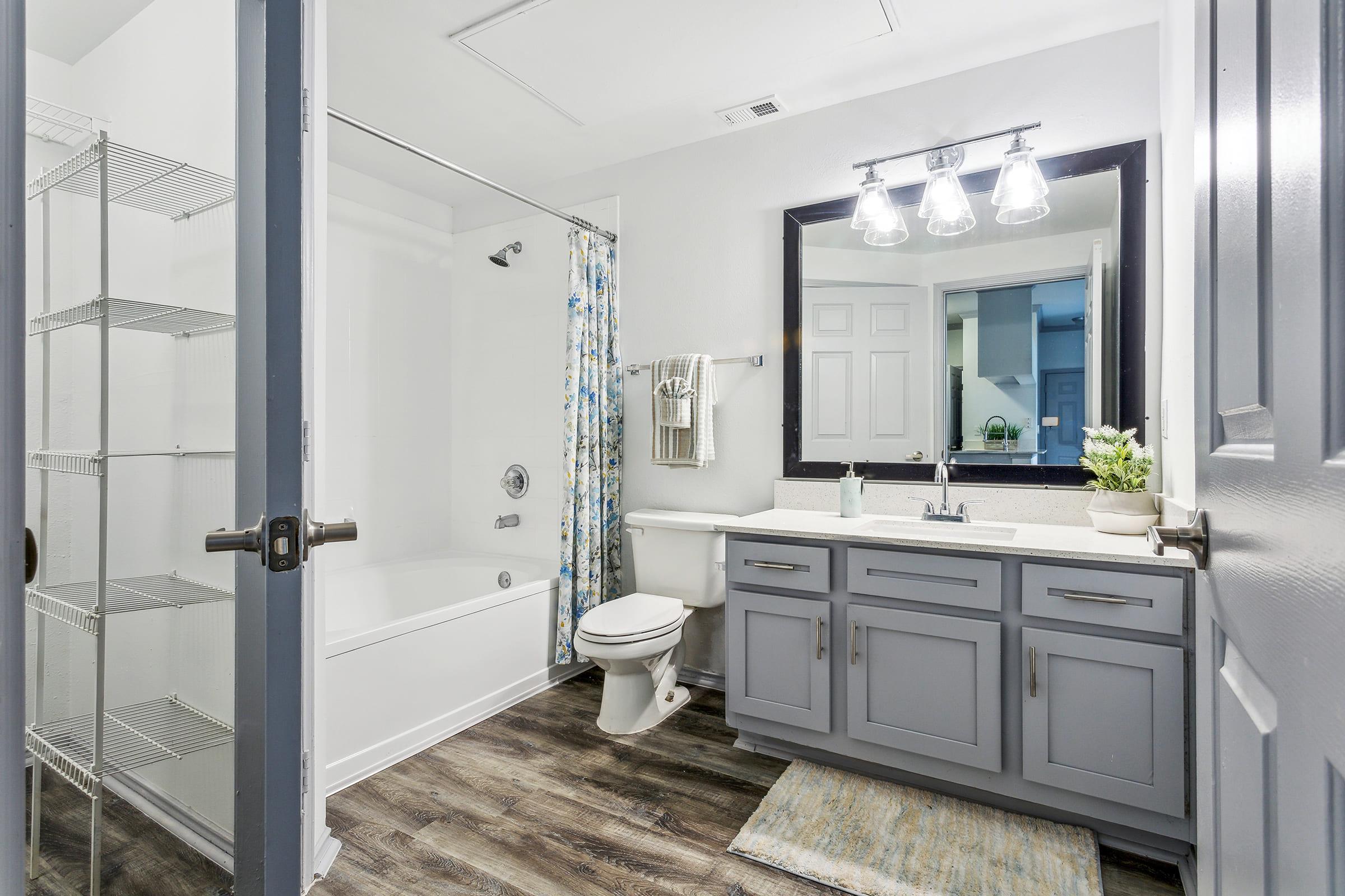 Modern bathroom featuring a bathtub with a shower curtain, a gray vanity with a sink, a toilet, and a mirror above the sink. The floor is finished with wooden laminate, and there are shelves for storage. Natural light enters through the door. A small plant adds a touch of decor.
