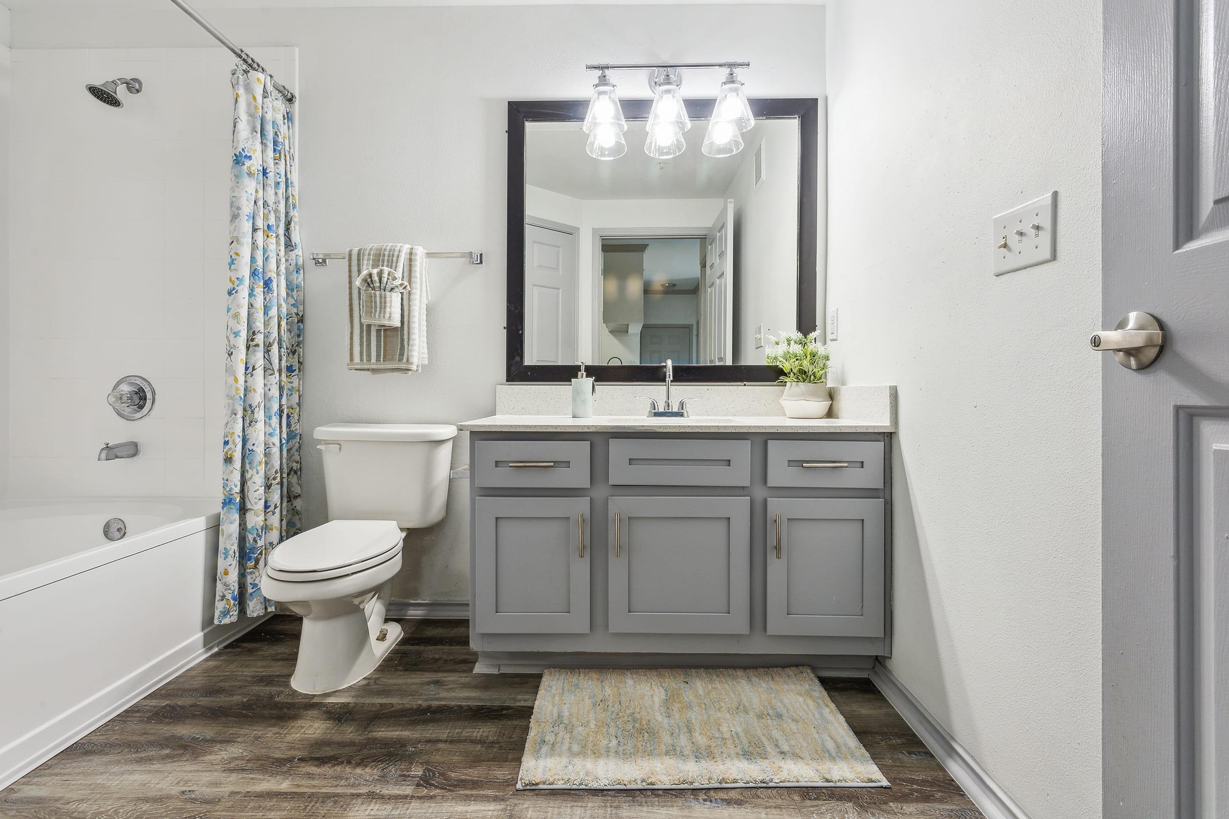 Modern bathroom featuring a shower/tub combo with a floral curtain, a gray vanity with a countertop sink, a mirror with three light fixtures above, and a gray door. The floor is wooden with a decorative bath mat. The walls are painted white, creating a bright and clean atmosphere.