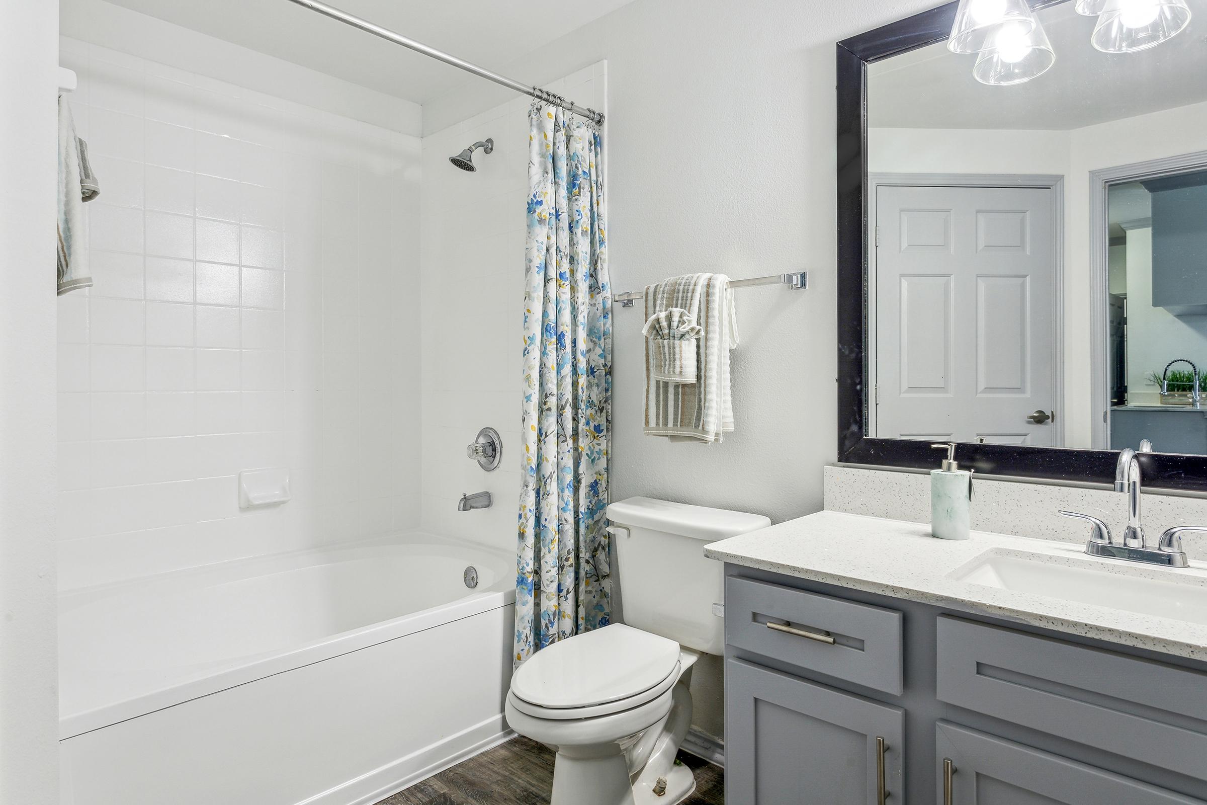 A clean and modern bathroom featuring a bathtub with a white shower curtain, a toilet, and a vanity with a sink. The walls are painted light gray, and there is a large mirror above the sink. A blue floral shower curtain adds a pop of color to the space. Soft lighting illuminates the room.