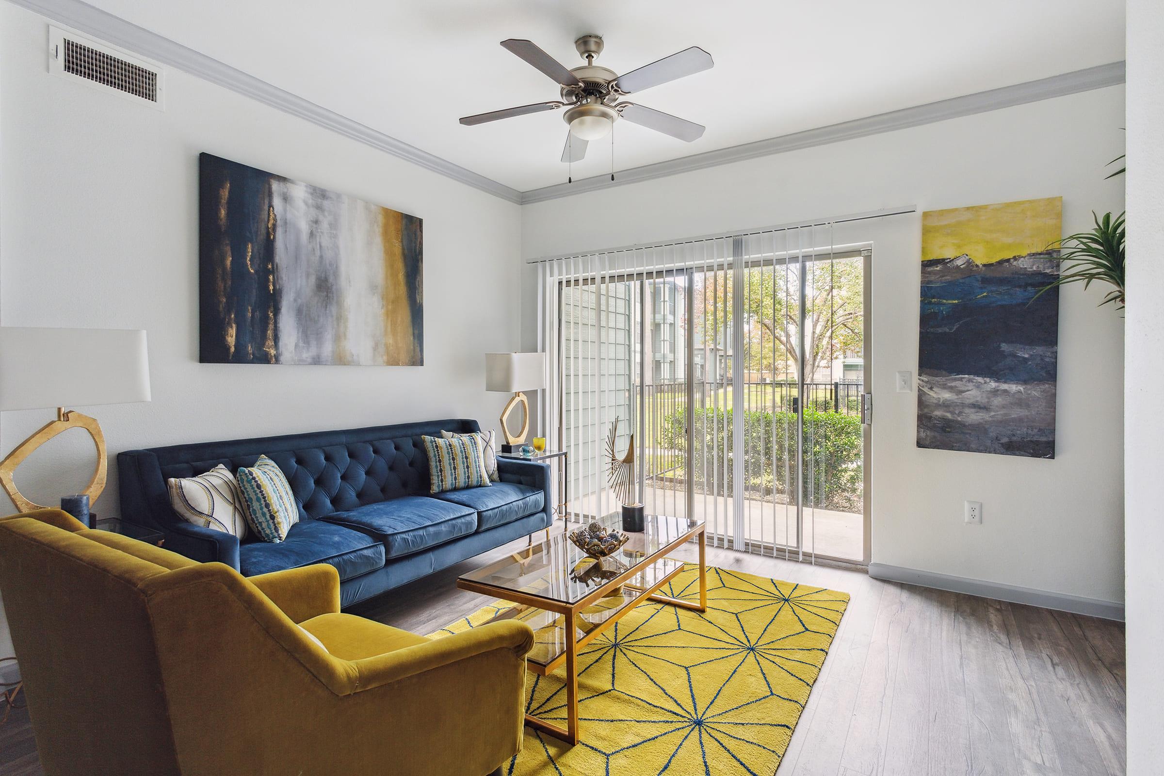 A cozy living room featuring a blue sofa and a yellow armchair. A glass coffee table sits on a yellow and blue patterned rug. Large windows with sheer curtains allow natural light in, while abstract paintings hang on the walls. Indoor plants add a touch of greenery to the space.