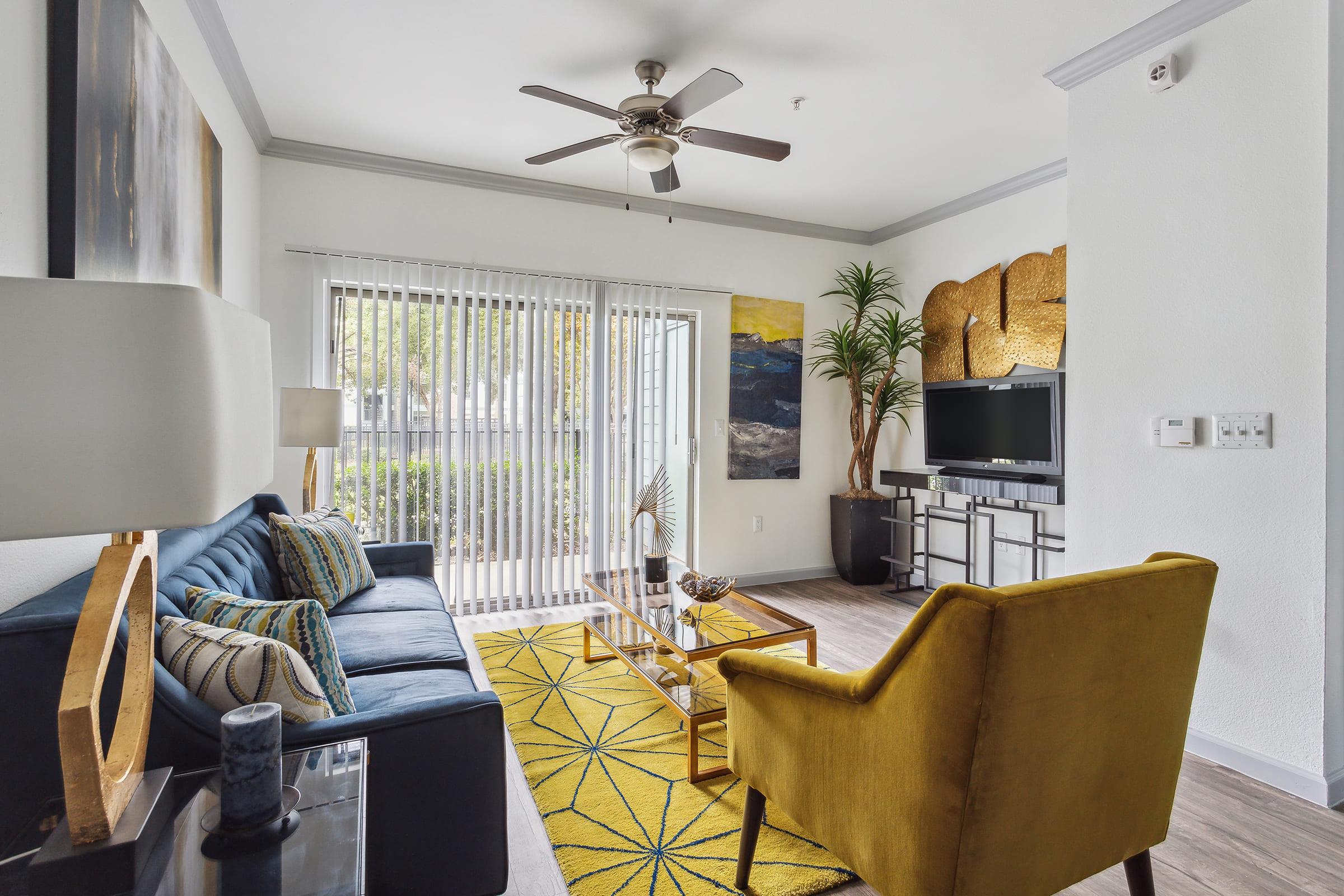 A modern living room featuring a blue sofa with patterned cushions, a yellow accent chair, and a glass coffee table on a geometric yellow rug. There’s a flat-screen TV on a media console, large windows with blinds, and decorative artwork on the walls, creating a bright and inviting atmosphere.