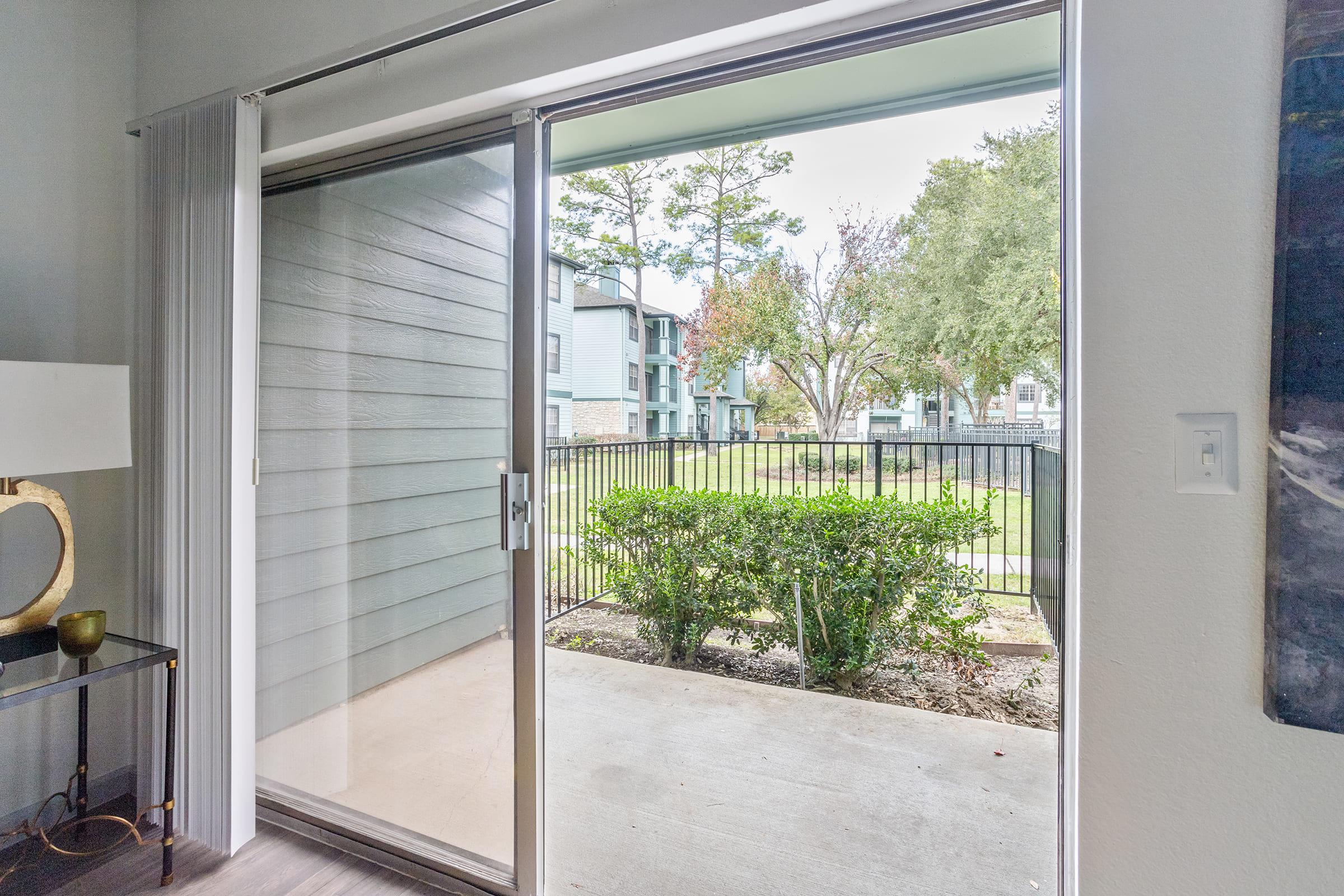 A view from inside an apartment looking out through a sliding glass door onto a small patio and a grassy area. Trees and apartment buildings are visible in the background, indicating a residential community. The door is open, allowing natural light to enter the indoor space.