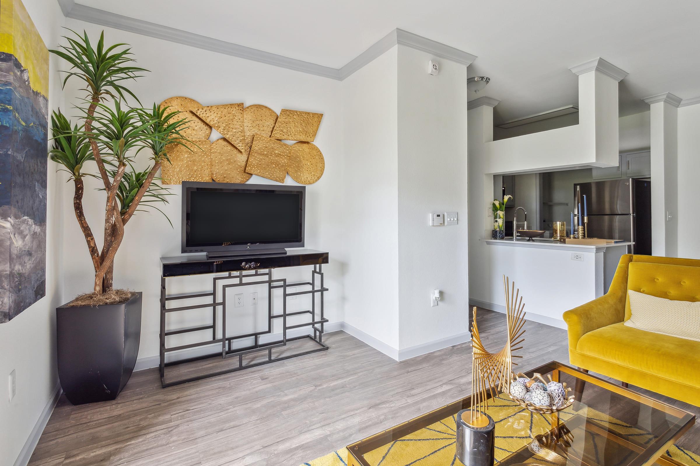 A modern living room with a yellow accent chair, a sleek black TV stand, and decorative wall art. A potted plant adds greenery, while a coffee table featuring a geometric design and decorative objects contributes to the contemporary aesthetic. The kitchen is visible in the background.