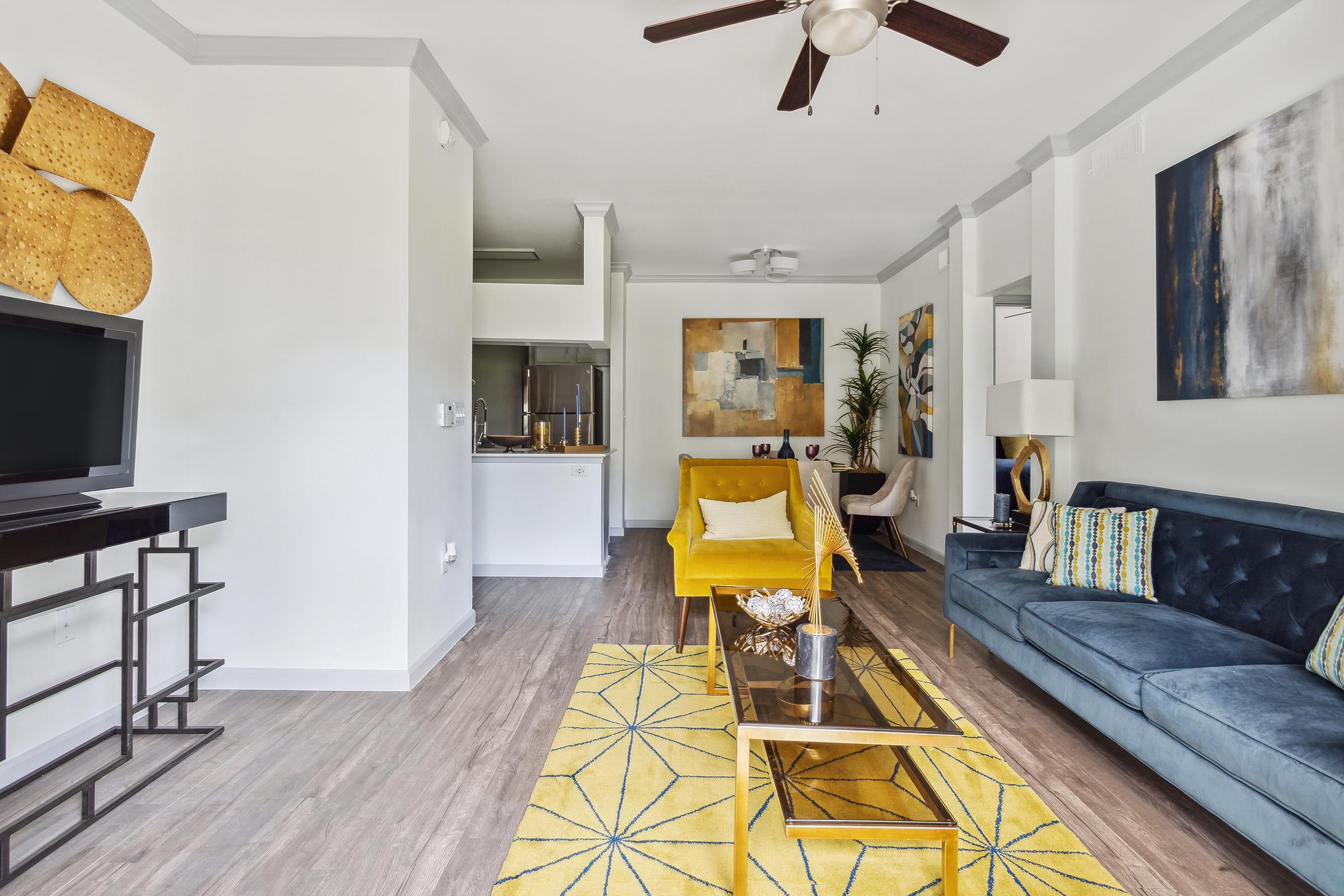 A modern living room featuring a blue sofa, a yellow armchair, and a decorative coffee table with a gold frame. The walls are adorned with abstract art, and a yellow geometric rug covers the floor. A fan hangs from the ceiling, and the space is brightened by natural light. There are plants and stylish decor accents throughout.