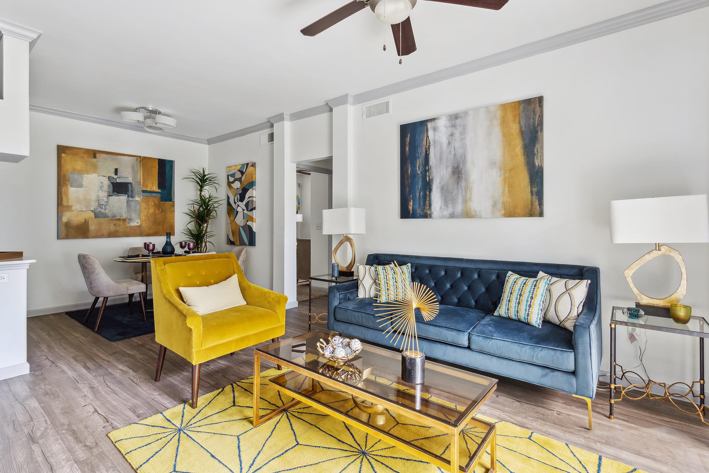 A modern living room featuring a blue tufted sofa and a bright yellow accent chair. The space includes a glass coffee table on a geometric patterned rug, decorative lamps, and abstract artwork on the walls. A potted plant adds a touch of greenery, creating a stylish, welcoming atmosphere.