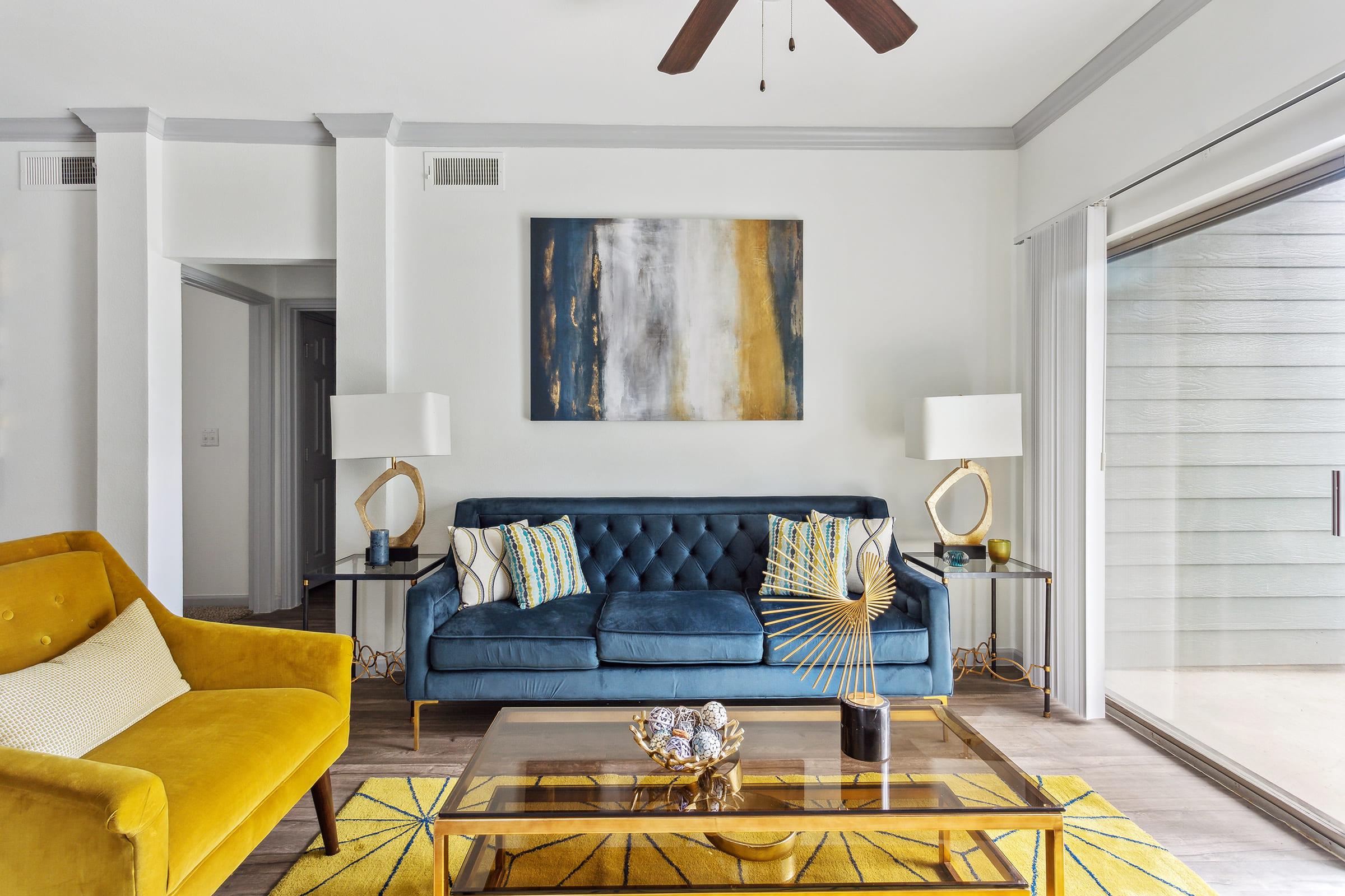 A stylish living room featuring a blue tufted sofa with decorative pillows, a yellow accent chair, a glass coffee table with a decorative centerpiece, and a large abstract painting on the wall. The space is bright with natural light coming through a sliding glass door, complementing the modern decor.