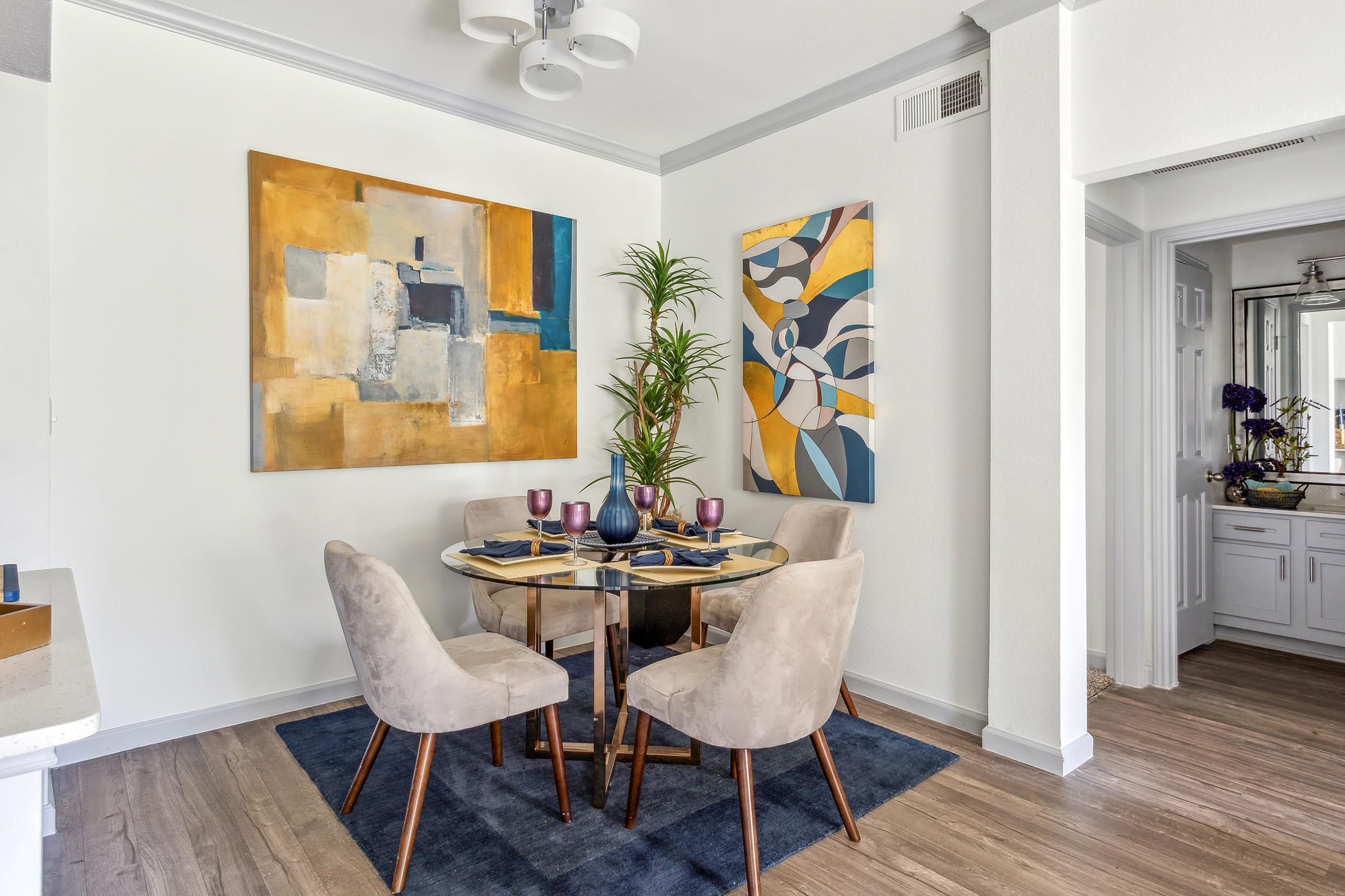 A modern dining area featuring a round glass table surrounded by four beige upholstered chairs. The walls are adorned with two abstract paintings, one in warm tones and the other with blue and yellow hues. A green plant adds a touch of nature, and a small piece of furniture is visible in the background.