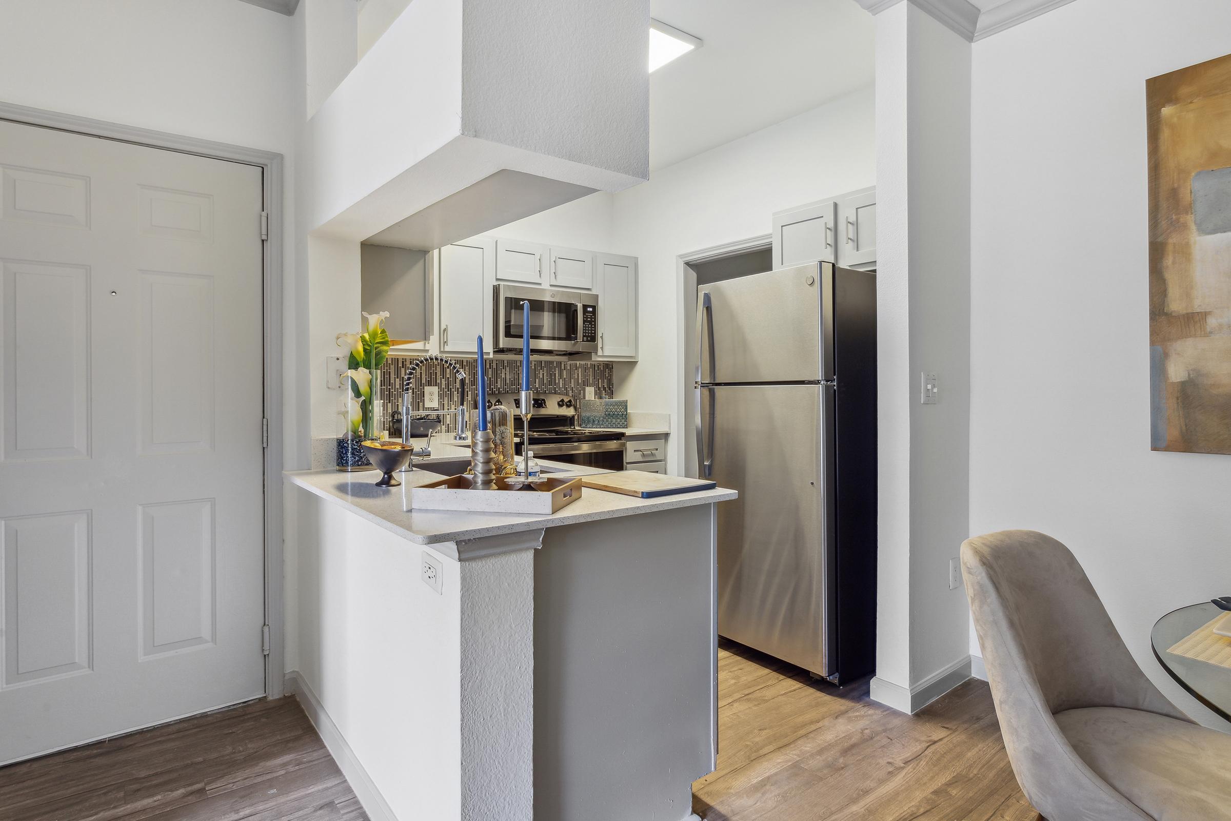 A modern kitchen view featuring an open layout. The image shows a doorway leading into the kitchen, with stainless steel appliances, white cabinets, and a countertop displaying a decorative arrangement. Natural light illuminates the space, enhancing the inviting atmosphere.