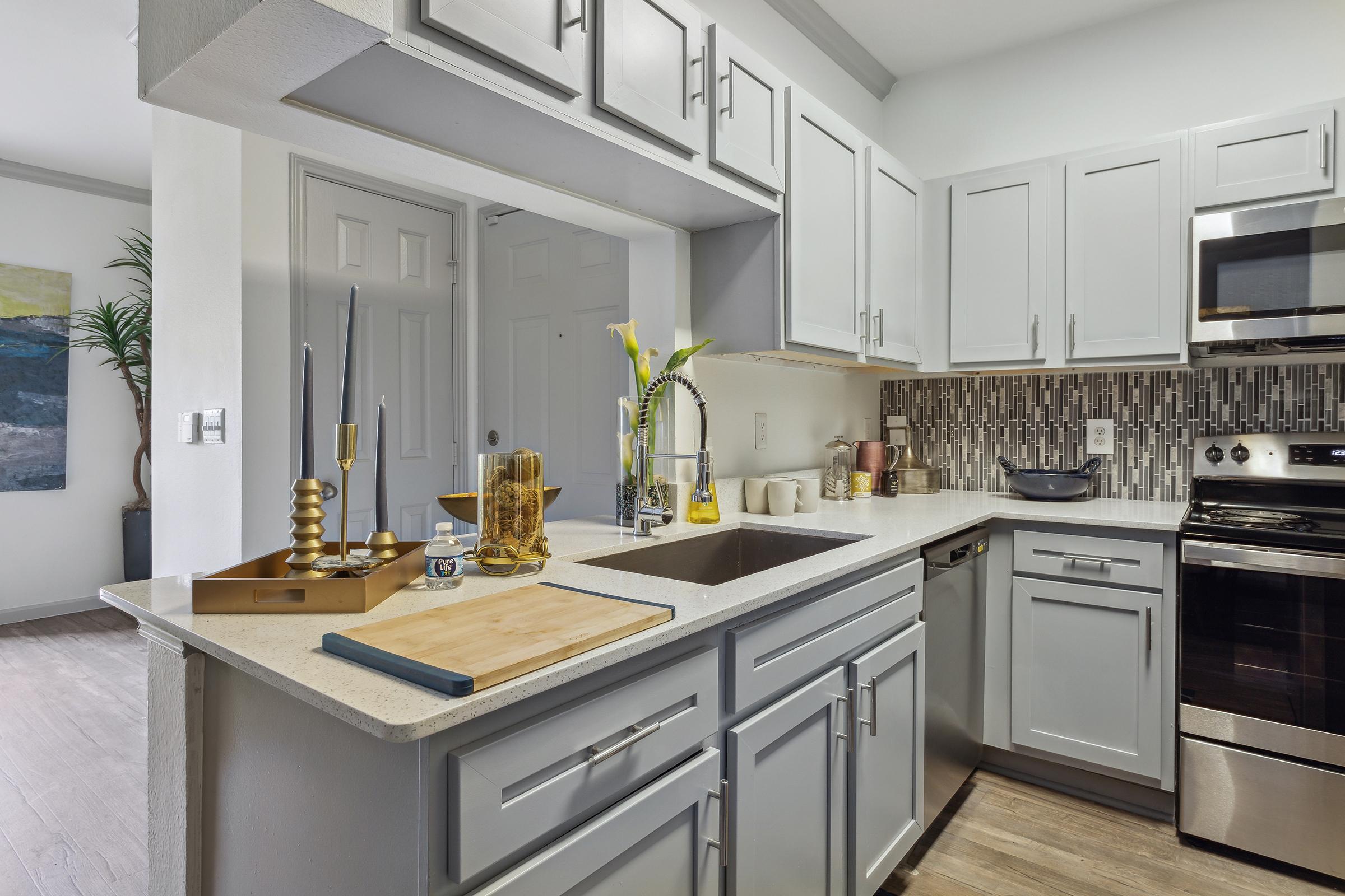 Modern kitchen with light gray cabinets, a stainless steel stove, and a sleek countertop. Features include a cutting board, decorative items, and a backsplash with mosaic tiles. A plant is visible in the background, contributing to the contemporary aesthetic. Well-lit and organized space.