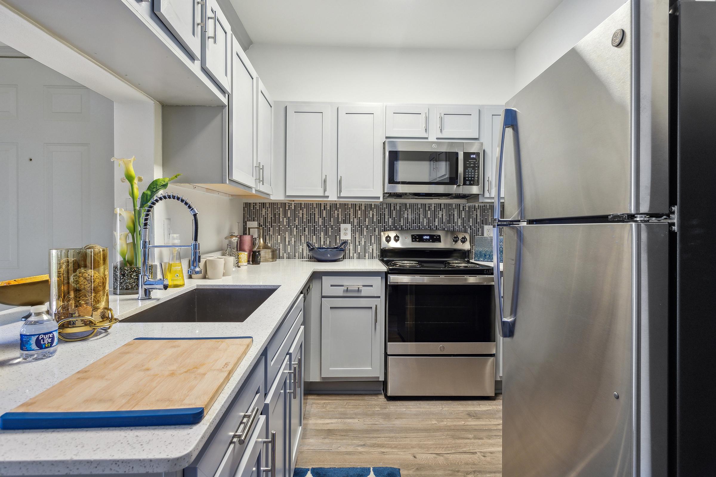 A modern kitchen featuring gray cabinets, a stainless steel refrigerator, an electric stove, and a microwave. The countertop is adorned with a cutting board, a vase with flowers, and various kitchen utensils. The backsplash consists of decorative tiles, and there's a blue rug on the floor.