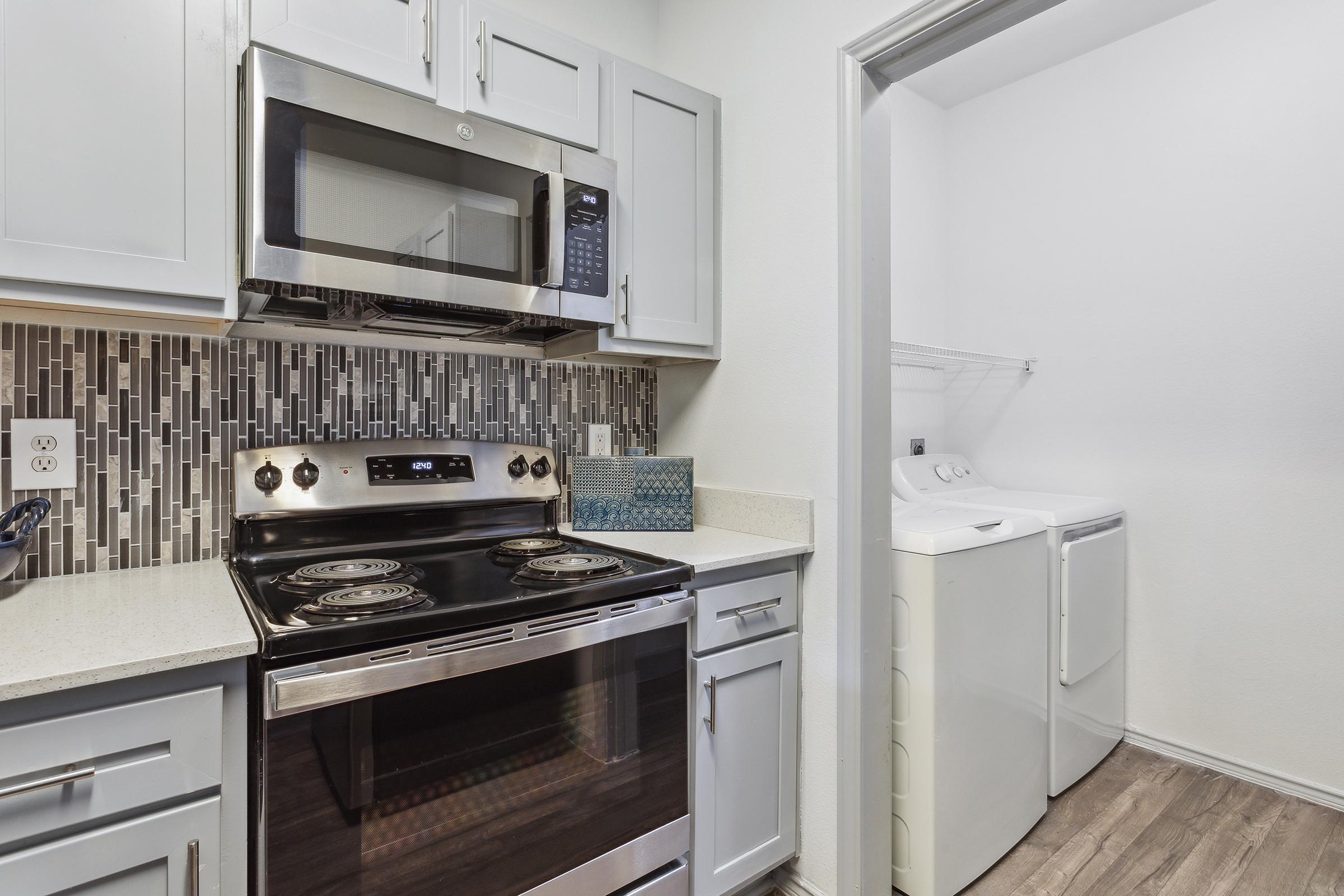 A modern kitchen featuring stainless steel appliances, including a microwave and an oven with a glass top stove. The cabinets are light blue, and there is a decorative backsplash of gray tiles. To the side, a laundry area is visible with a washer and dryer, and the flooring is a warm wood tone.