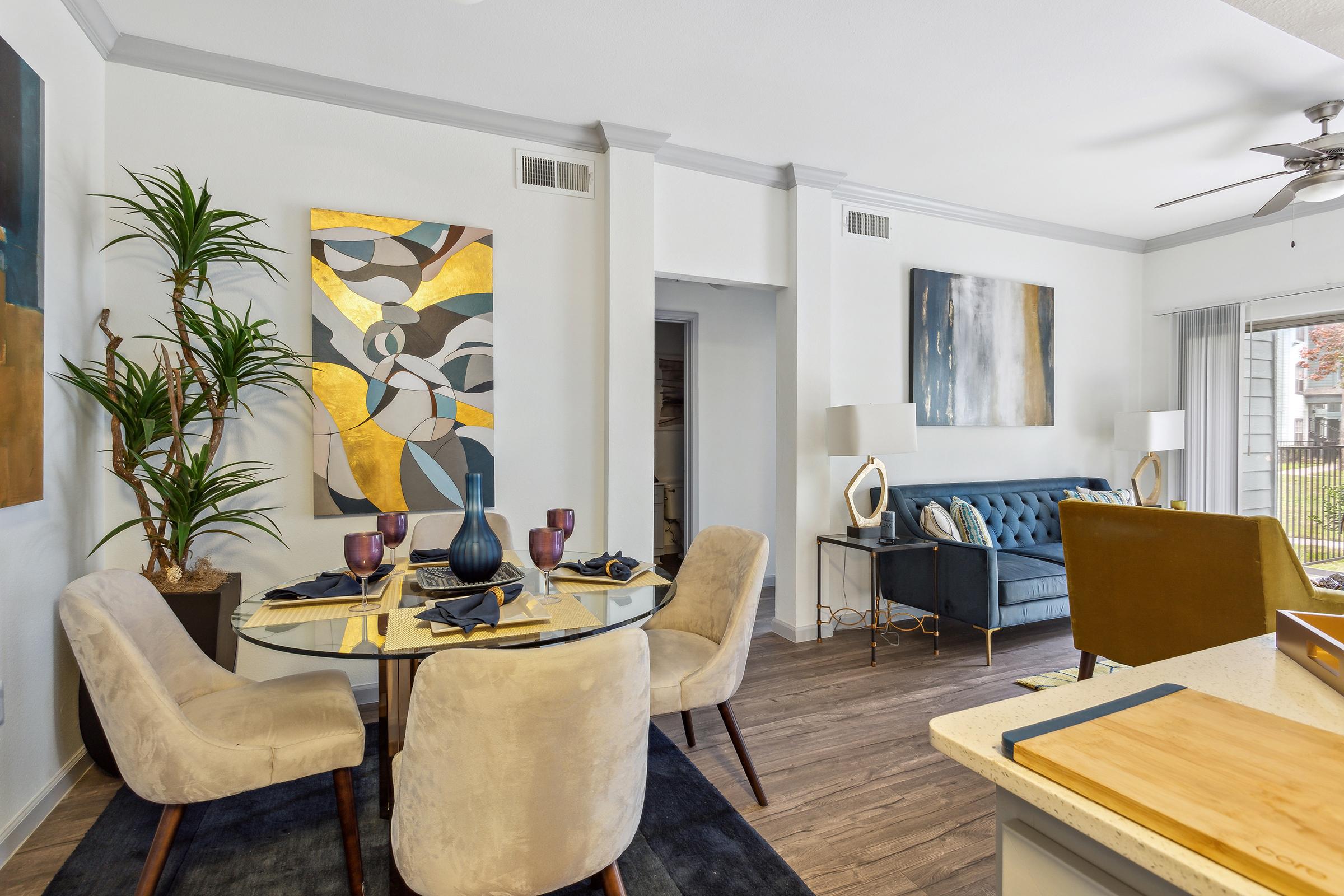A modern living space featuring a round dining table set for four with purple glasses, surrounded by upholstered chairs. The decor includes abstract artwork on the walls and a cozy blue sofa with cushions. A large window allows natural light, and potted plants add greenery to the room.