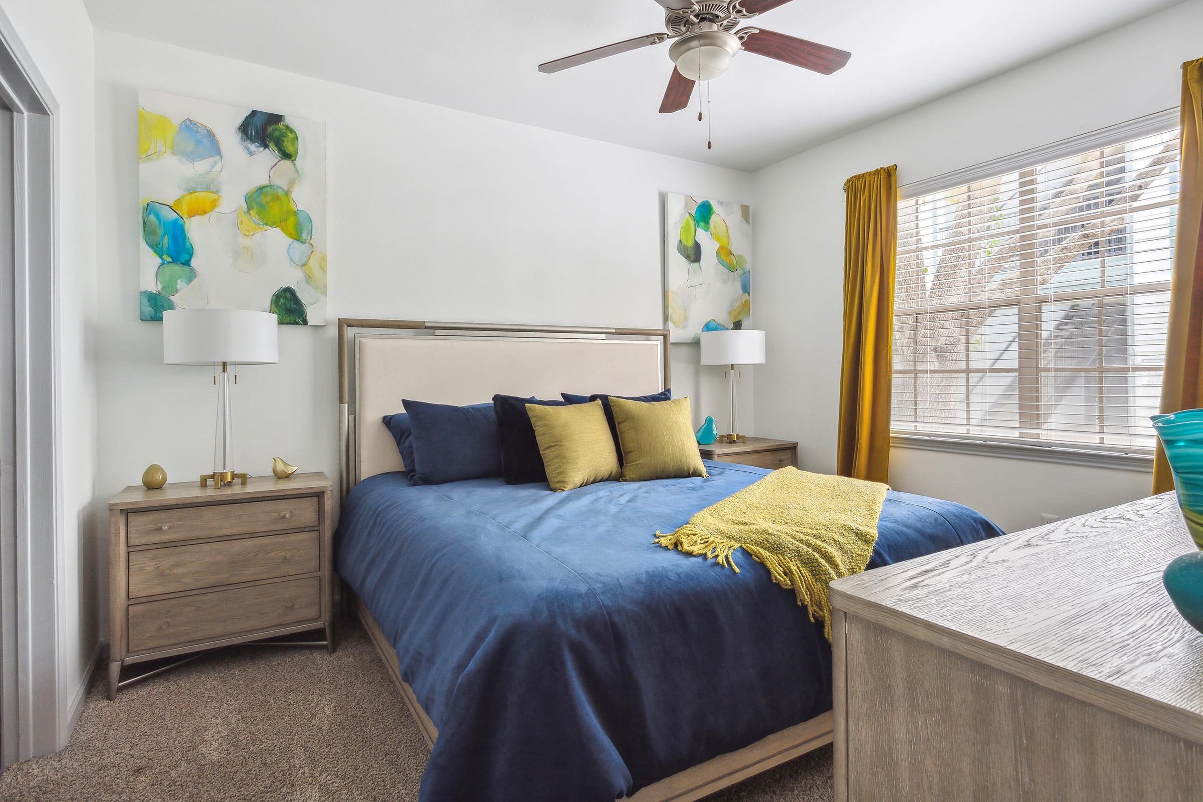 A well-decorated bedroom featuring a blue bedspread with pillows, yellow accents, and wooden furniture. The walls are adorned with abstract art pieces in green and blue tones. Natural light filters through a window with yellow curtains, creating a cozy atmosphere.