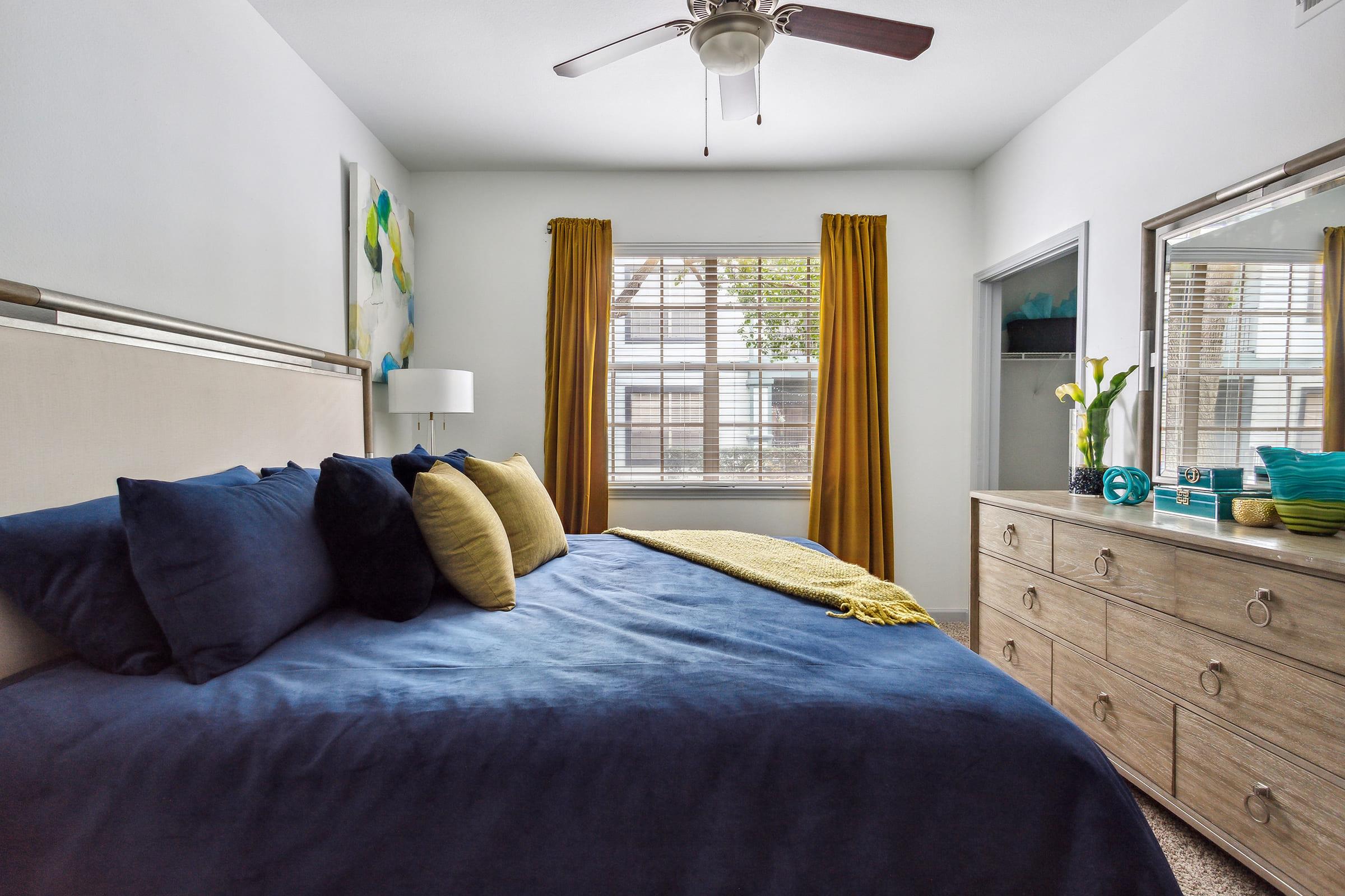 A cozy bedroom featuring a large bed with navy blue bedding and colorful accent pillows. Light yellow curtains frame a window, allowing natural light in. A dresser with a decorative vase sits nearby, and a ceiling fan adds a modern touch to the space.