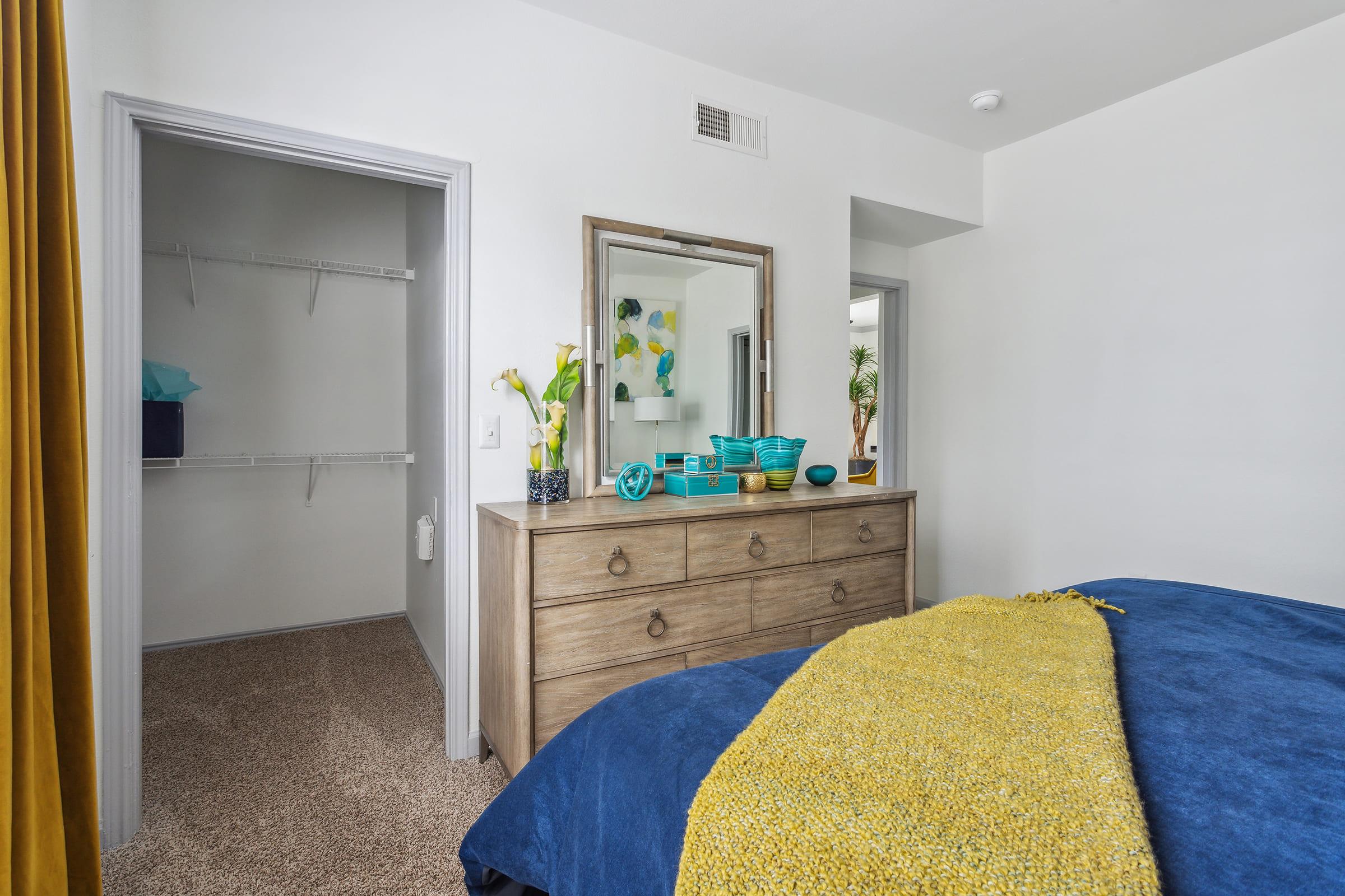 A cozy bedroom featuring a large, upholstered bed with a blue blanket and a bright yellow throw. A wooden dresser with a mirror displays decorative vases and bowls. A closet is visible to the left, and the walls are painted white, creating a fresh and airy atmosphere.