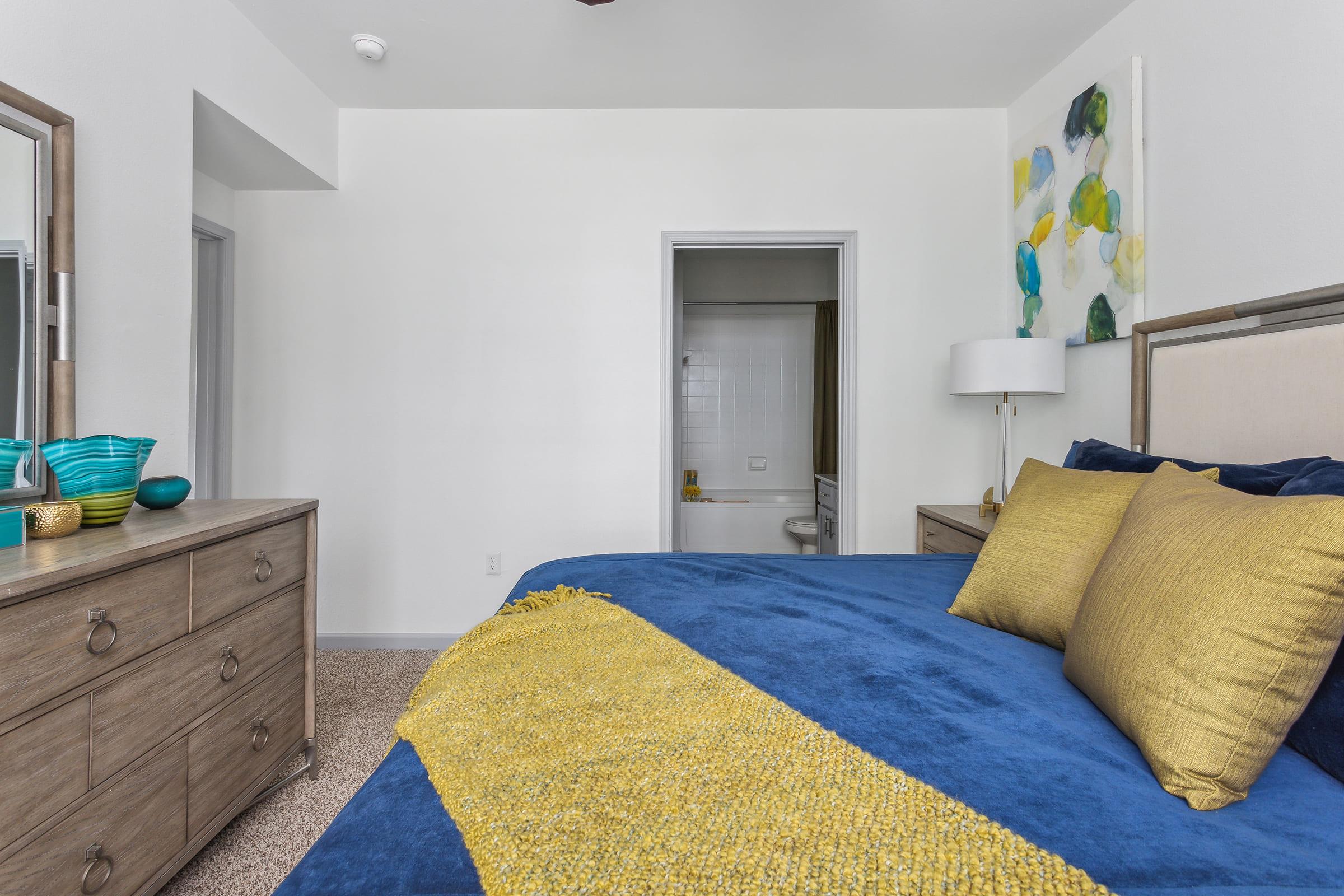 A modern bedroom featuring a blue bedspread with yellow accent pillows, a wooden dresser with decorative items, and a framed artwork on the wall. A door leads to a bathroom, and there's a lamp on a bedside table, creating a bright and inviting atmosphere.