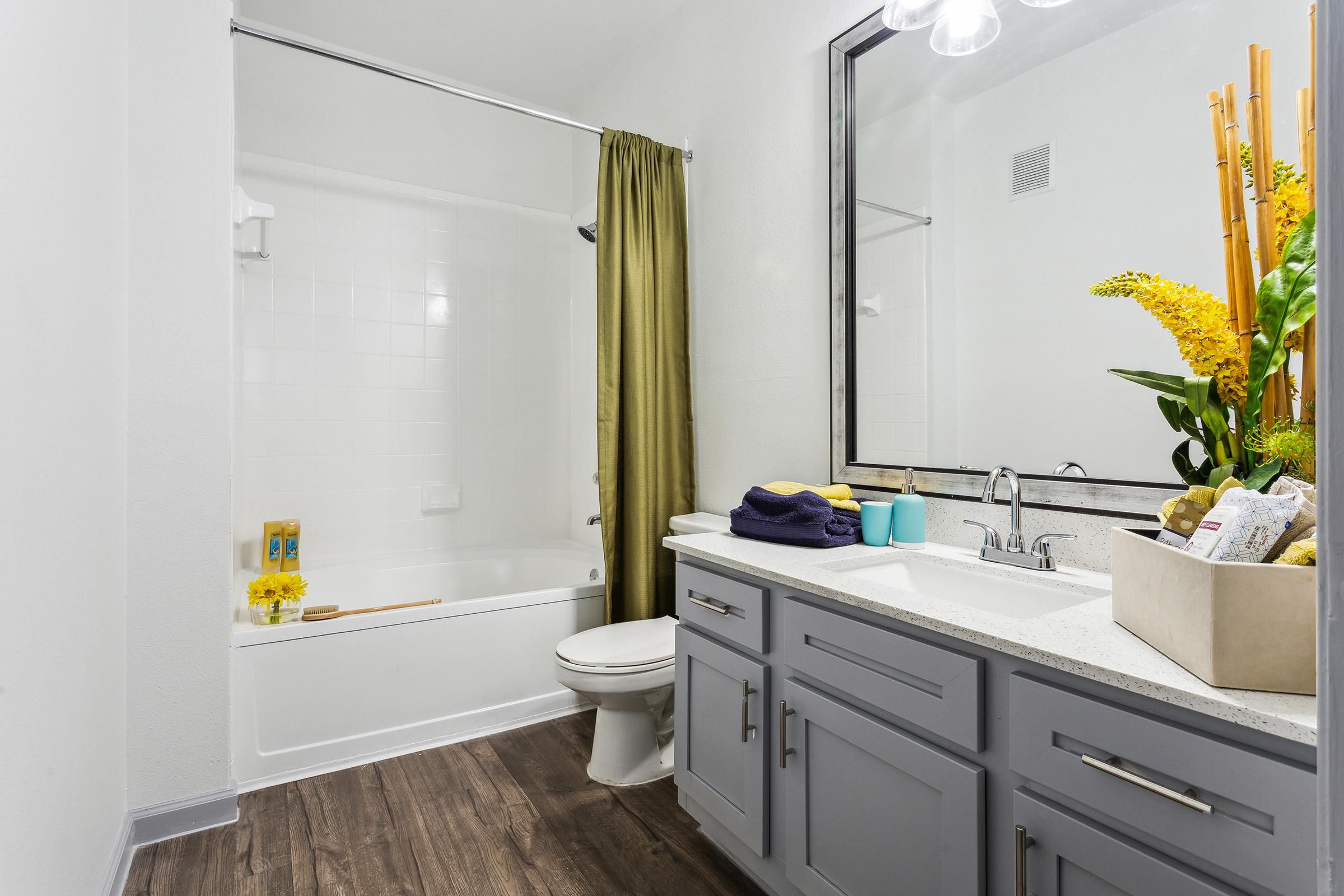A modern bathroom featuring a white bathtub with a shower curtain, a gray vanity with a marble countertop, a large mirror, and decorative plants. The decor includes yellow flowers and towels neatly arranged, creating a clean and inviting atmosphere.