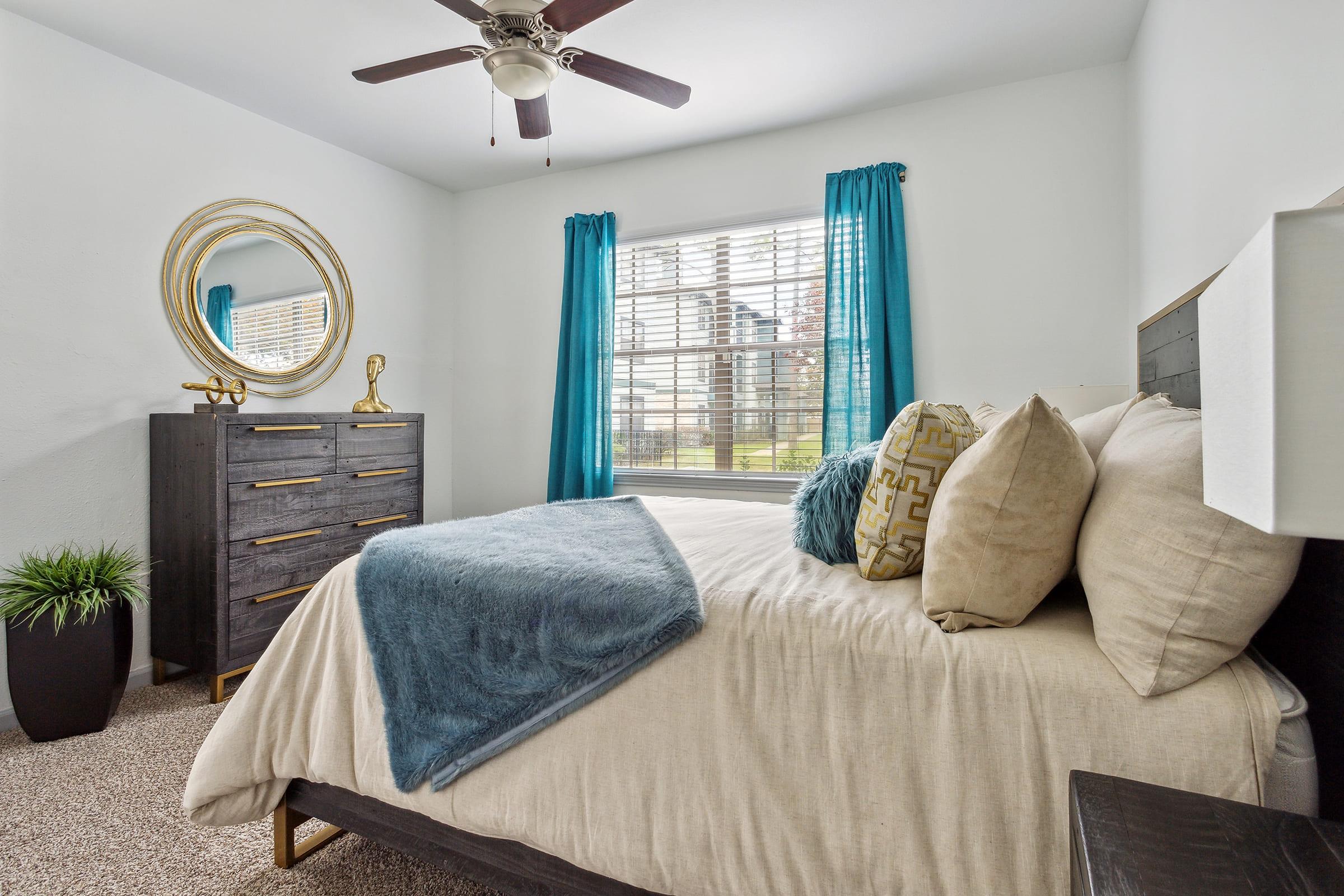 A cozy bedroom featuring a large window with teal curtains, a neatly made bed with decorative pillows and a soft throw blanket. A stylish dresser with a round mirror sits opposite the bed, and a small potted plant adds a touch of greenery to the space. The walls are painted white, creating a bright atmosphere.