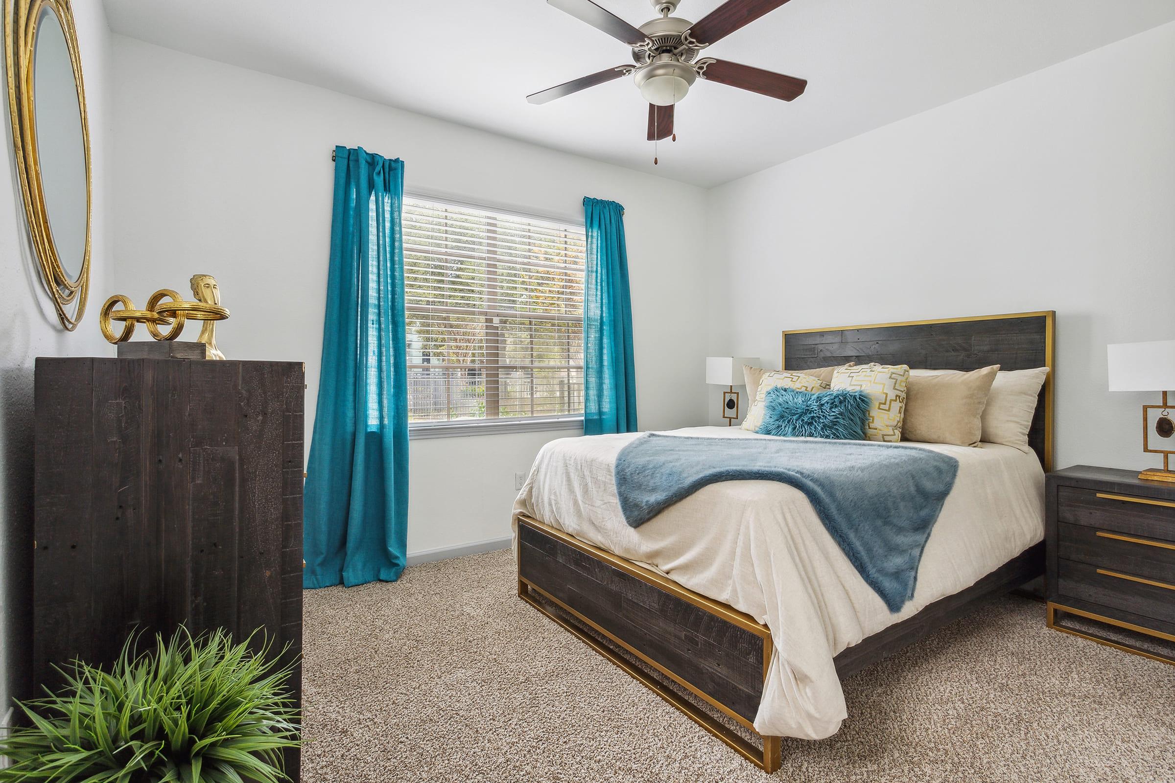 A cozy bedroom featuring a queen-sized bed with a white and blue bedspread, decorative pillows, and a wooden headboard. There's a modern nightstand and dresser, along with a round gold-framed mirror on the wall. Bright blue curtains frame a window, allowing natural light to illuminate the space, complemented by a potted plant in the corner.