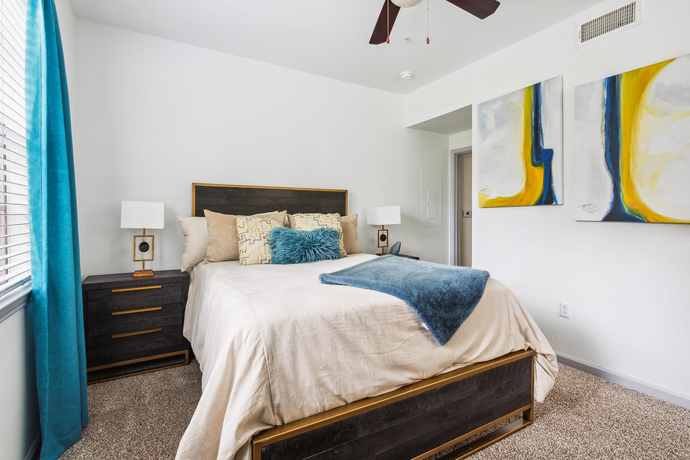 Cozy bedroom featuring a queen-sized bed with light bedding and a blue throw blanket. There are two stylish nightstands with lamps on either side of the bed. The room has large windows with teal curtains and two abstract paintings on the wall, creating a modern and inviting atmosphere.
