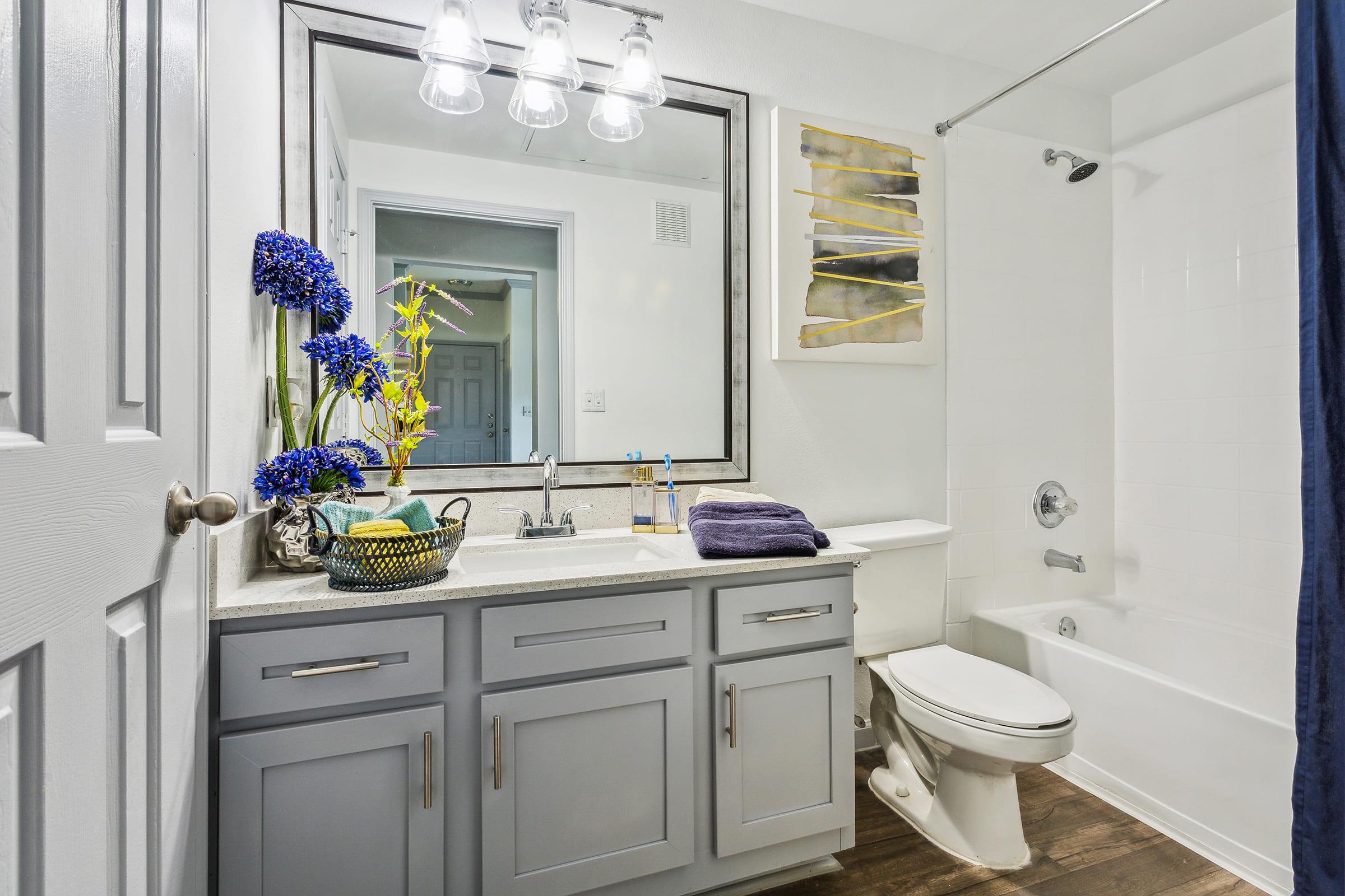 A modern bathroom featuring a light grey vanity with a sink, a large mirror, and decorative flowers. There's a bathtub with a shower curtain, a toilet, and wood-style flooring. Shelves display toiletries, and the overall ambiance is clean and inviting.