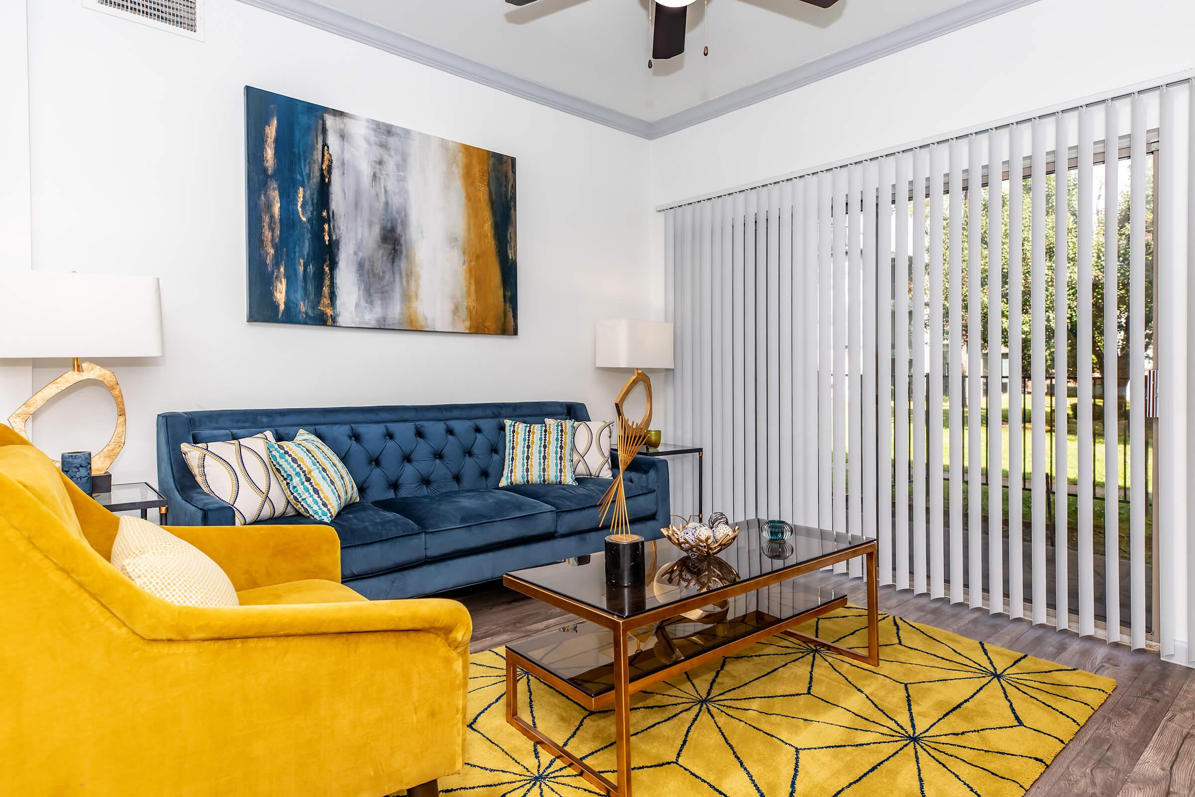 A stylish living room featuring a blue tufted sofa with decorative pillows, a yellow armchair, a glass coffee table with decorative items, and a vibrant yellow and gray area rug. The space has tall white blinds covering a large window, allowing natural light, and is adorned with modern wall art.