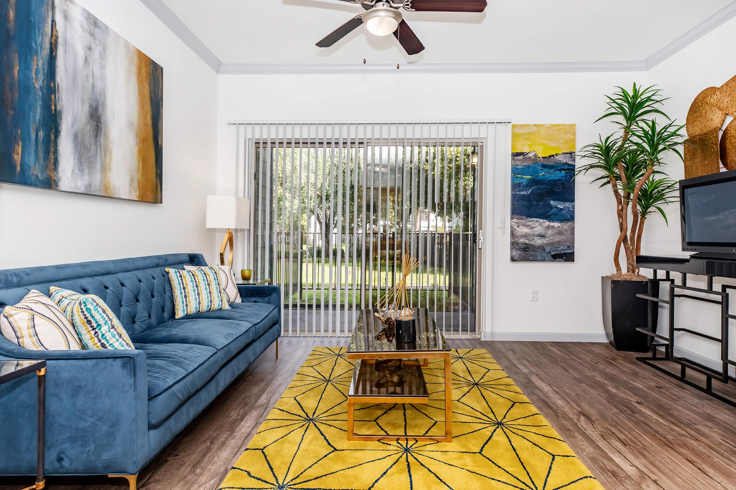 Modern living room featuring a blue sofa with decorative cushions, a glass coffee table on a yellow geometric-patterned rug, and large windows with vertical blinds. A tall indoor plant and colorful artwork adorn the walls, creating a bright and inviting atmosphere.