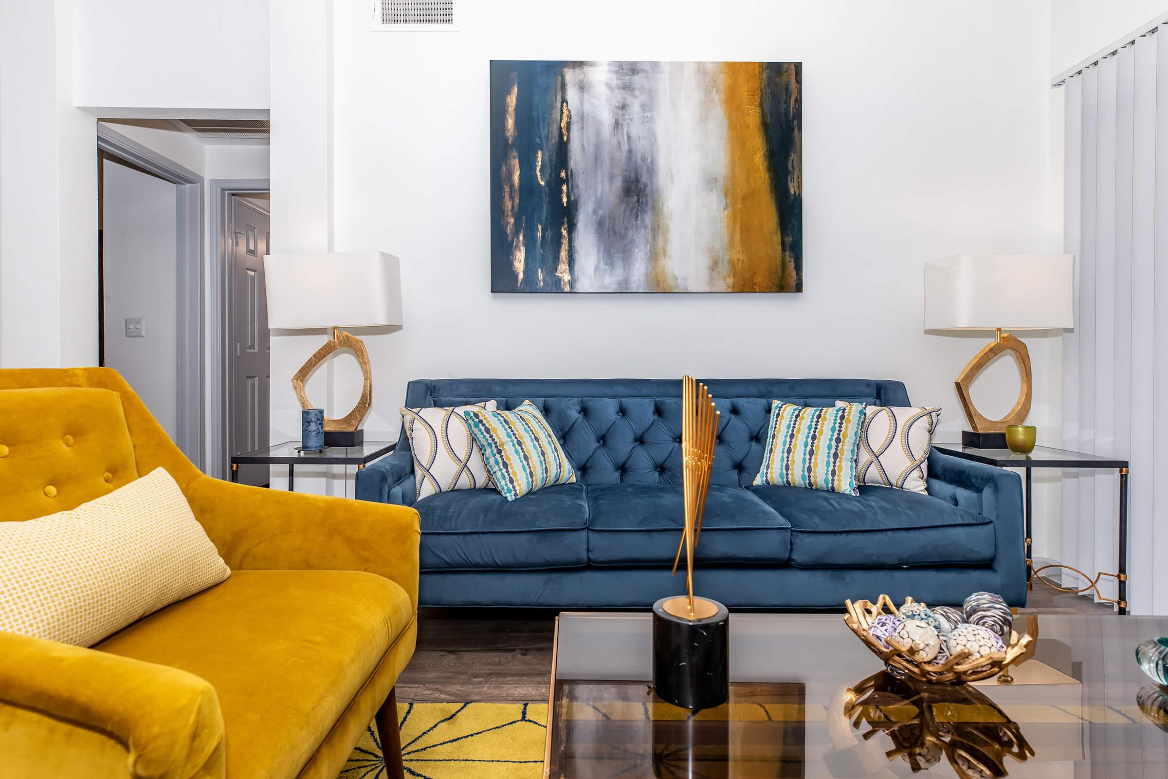 A cozy living room featuring a blue tufted sofa with decorative pillows, an elegant yellow armchair, a modern glass coffee table with decorative items, and two stylish table lamps. A large abstract painting with gold, blue, and white tones hangs on the wall, adding a contemporary touch to the space.