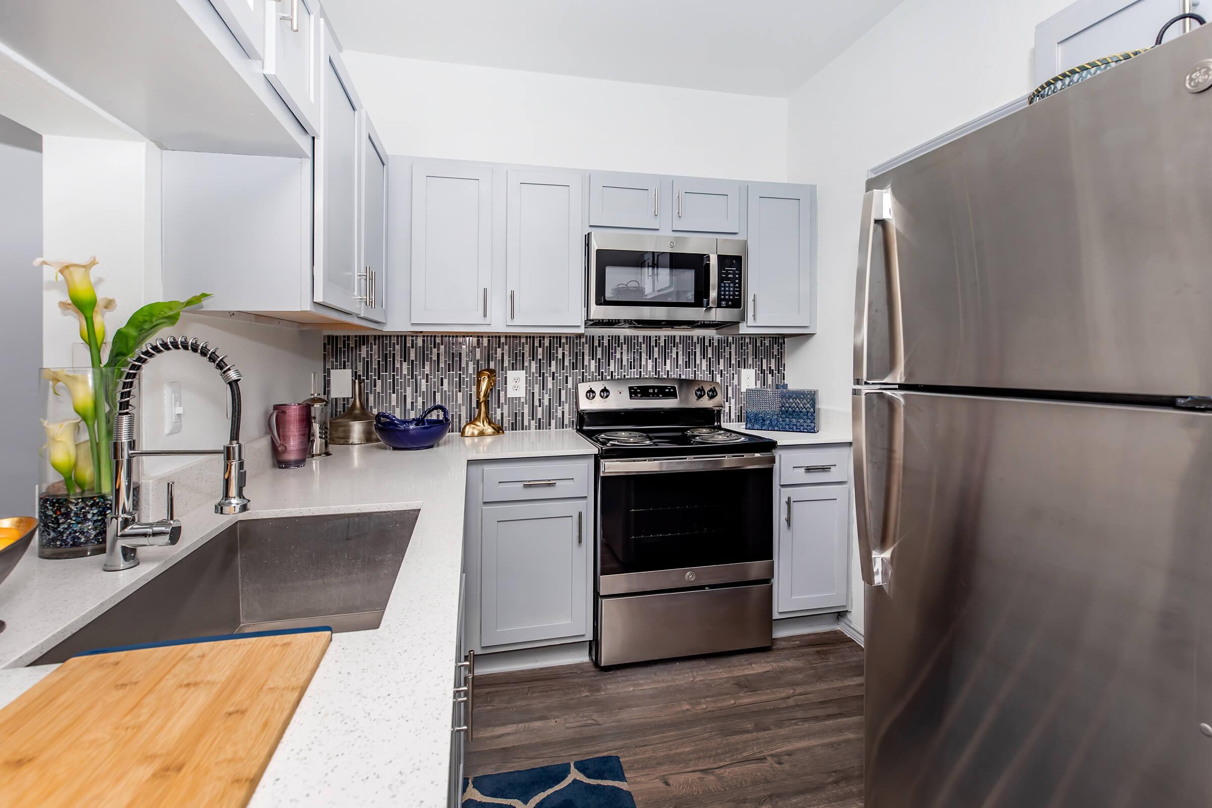 A modern kitchen featuring light gray cabinets, stainless steel appliances including a microwave and refrigerator, a double sink with a stylish faucet, and a countertop made of white stone. The back wall is adorned with decorative tiles, and a colorful area rug adds warmth to the space.