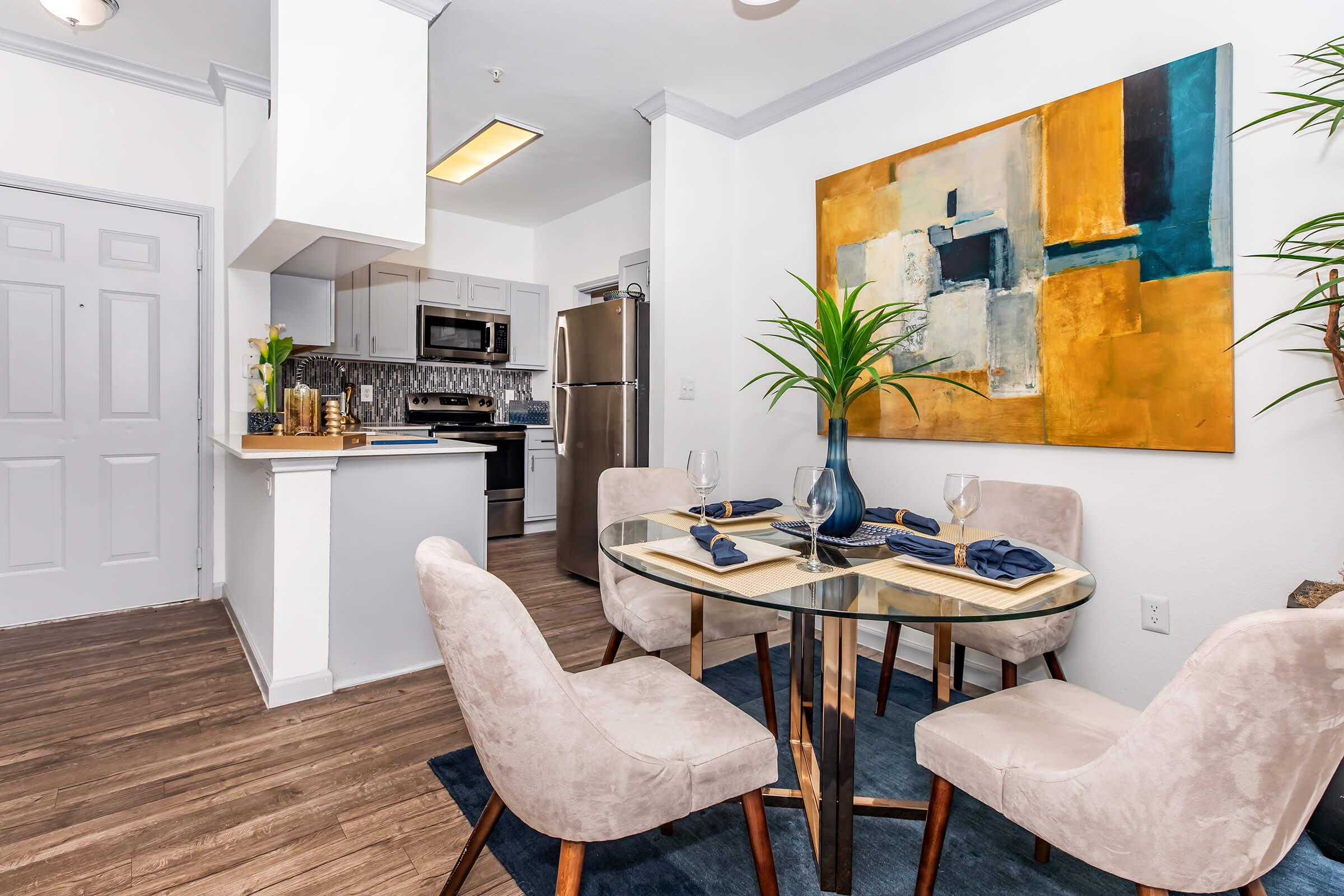 A modern kitchen and dining area featuring a glass table set for four, with plush chairs. The kitchen has stainless steel appliances and a backsplash. An abstract painting in warm tones hangs on the wall, complemented by greenery from a potted plant, creating a stylish and inviting atmosphere.