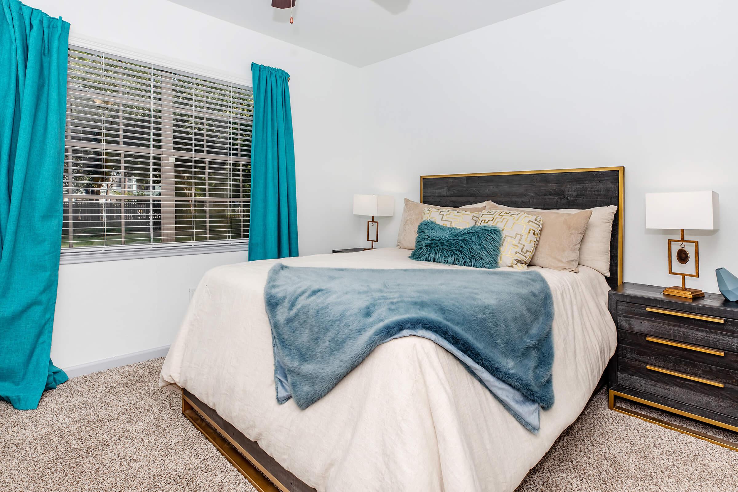 A cozy bedroom featuring a queen-sized bed with a light beige comforter and decorative pillows. There are two bedside tables with modern lamps, and large windows dressed in turquoise curtains allowing natural light. The room has a soft carpet, creating a warm and inviting atmosphere.