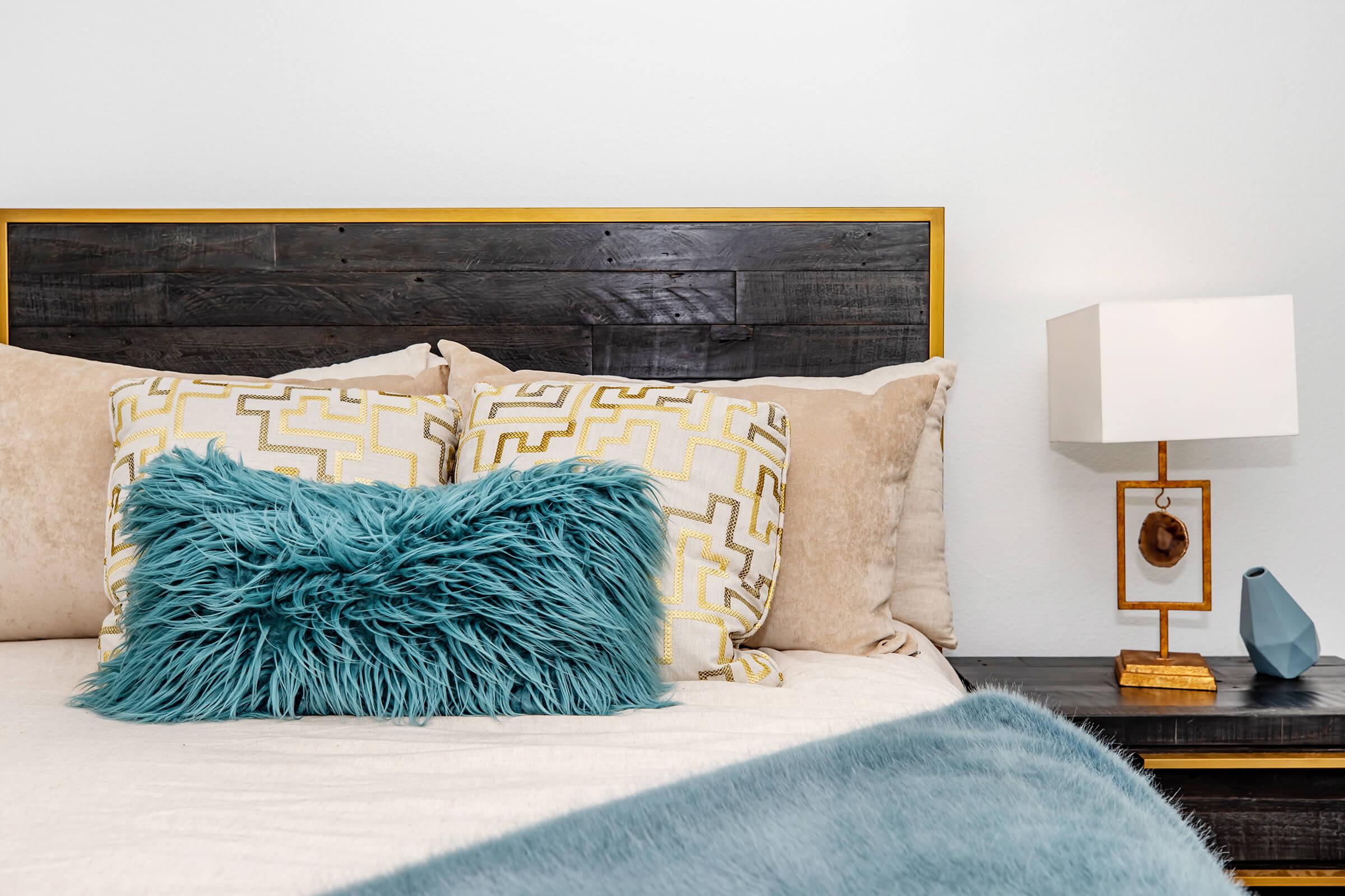 A cozy bedroom featuring a neatly made bed with decorative pillows, including a textured turquoise throw pillow. Next to the bed, there is a wooden nightstand with a modern lamp and a small decorative vase. The headboard has a rustic design with a warm color contrast.