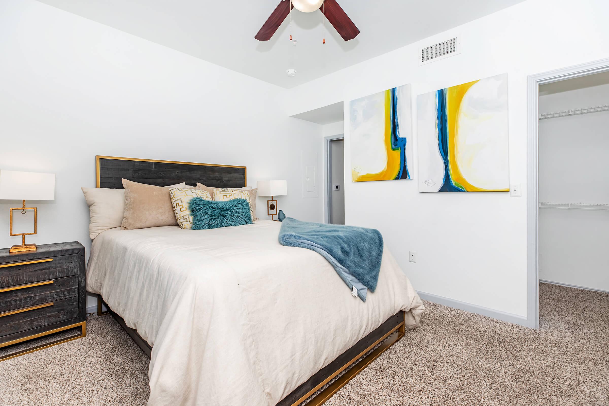 A cozy bedroom featuring a bed with a light-colored comforter and decorative pillows. There are two abstract paintings on the wall above the headboard, and a bedside table with a lamp. The room has soft lighting and a ceiling fan, with a closet visible in the background. The carpet is a neutral tone.