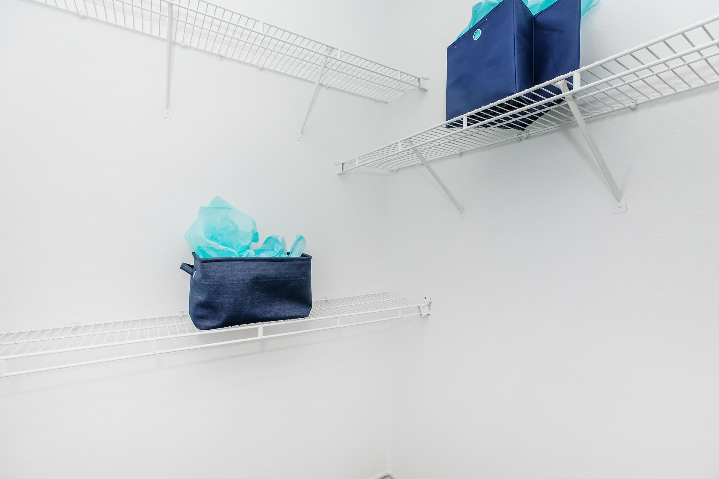 Empty storage space with white wire shelves. A dark blue storage bin with turquoise tissue paper sits on one of the shelves, while the other shelves remain unoccupied. The walls are plain white, creating a clean and organized appearance.