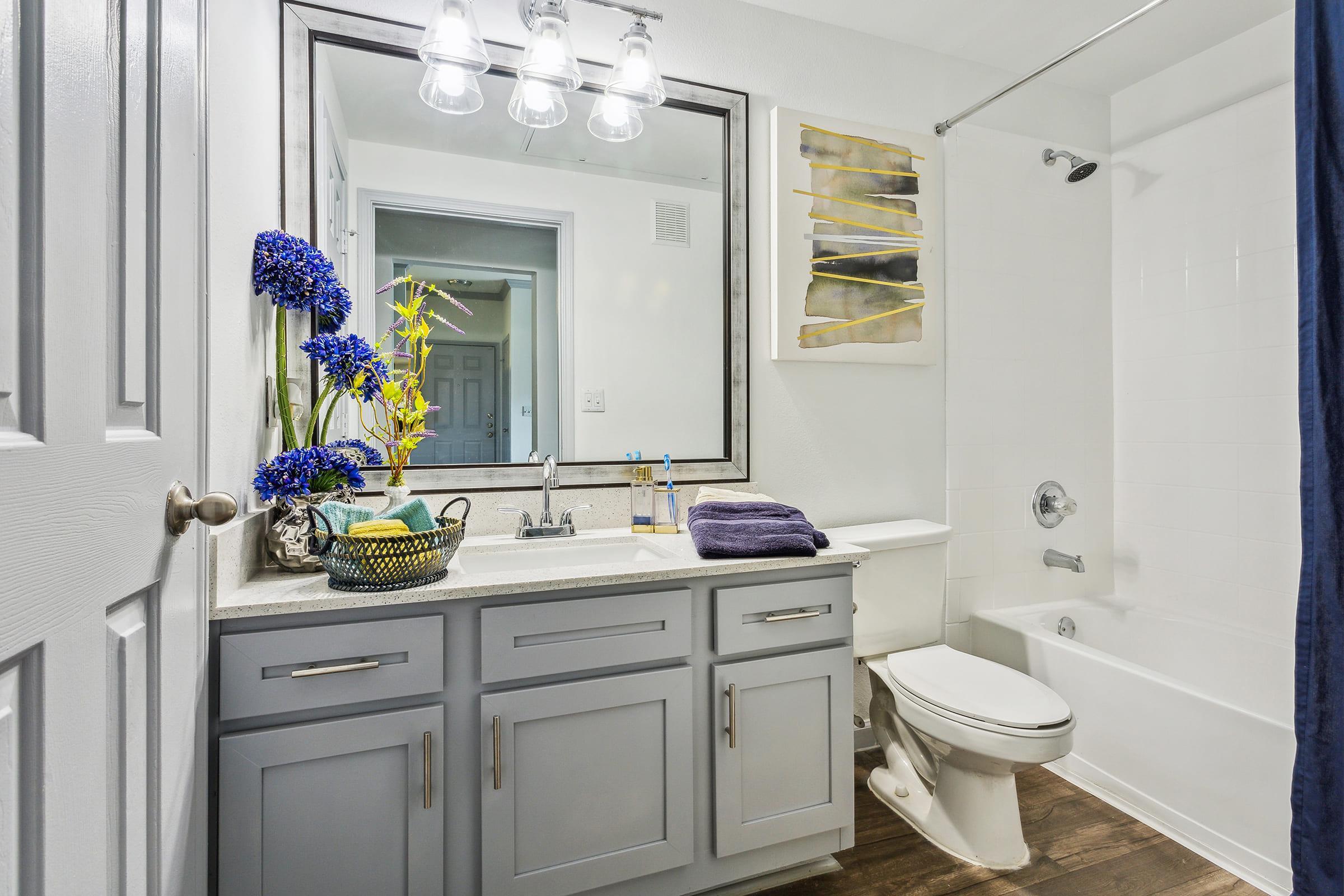 A modern bathroom featuring a light gray cabinet with a sink, a large mirror, and decorative blue flowers. The shower and bathtub area is white tiled, complemented by a navy blue curtain. The space is bright with natural light and includes organized toiletries on the counter.