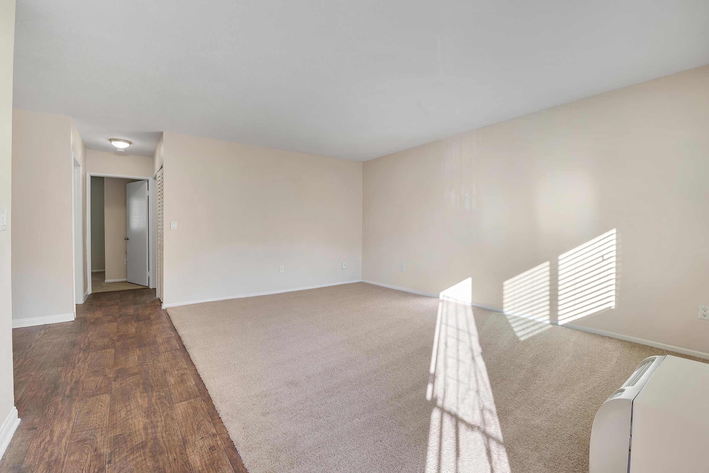 A spacious, empty room featuring light beige walls and light brown carpet. Sunlight filters in through a window, casting shadows on the wall. A small white heater is visible on the floor, and a doorway leads to another room in the back. The overall atmosphere is bright and inviting, ready for furnishings.