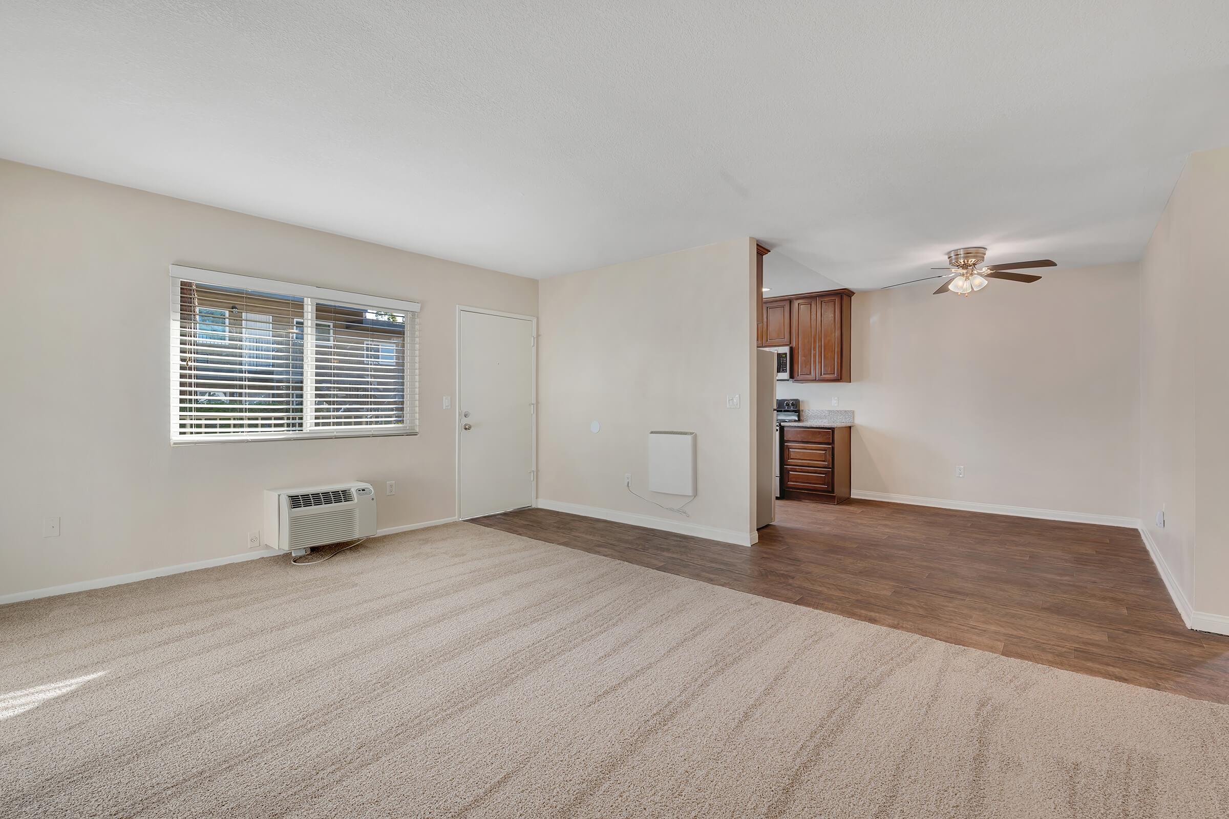 Spacious, neutral-toned living area featuring carpet flooring and large windows for natural light. There is a door leading outside, and a ceiling fan for ventilation. The kitchen is visible in the background with wooden cabinetry. Optional heating unit is located on the wall.