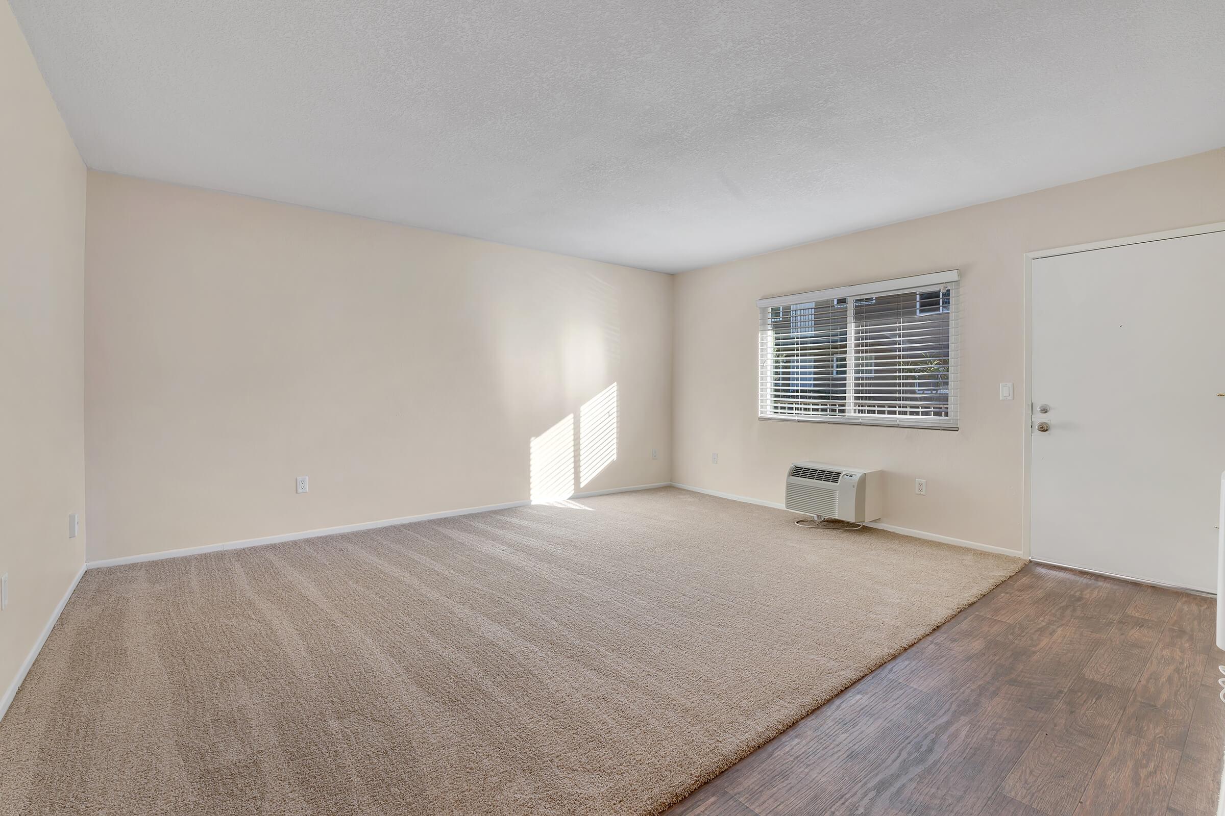 A spacious, empty room featuring light beige walls and a large window with horizontal blinds. The floor is covered with light-colored carpet, and there is a small air conditioning unit and a door leading outside. Natural light streams in, creating a bright and inviting atmosphere.