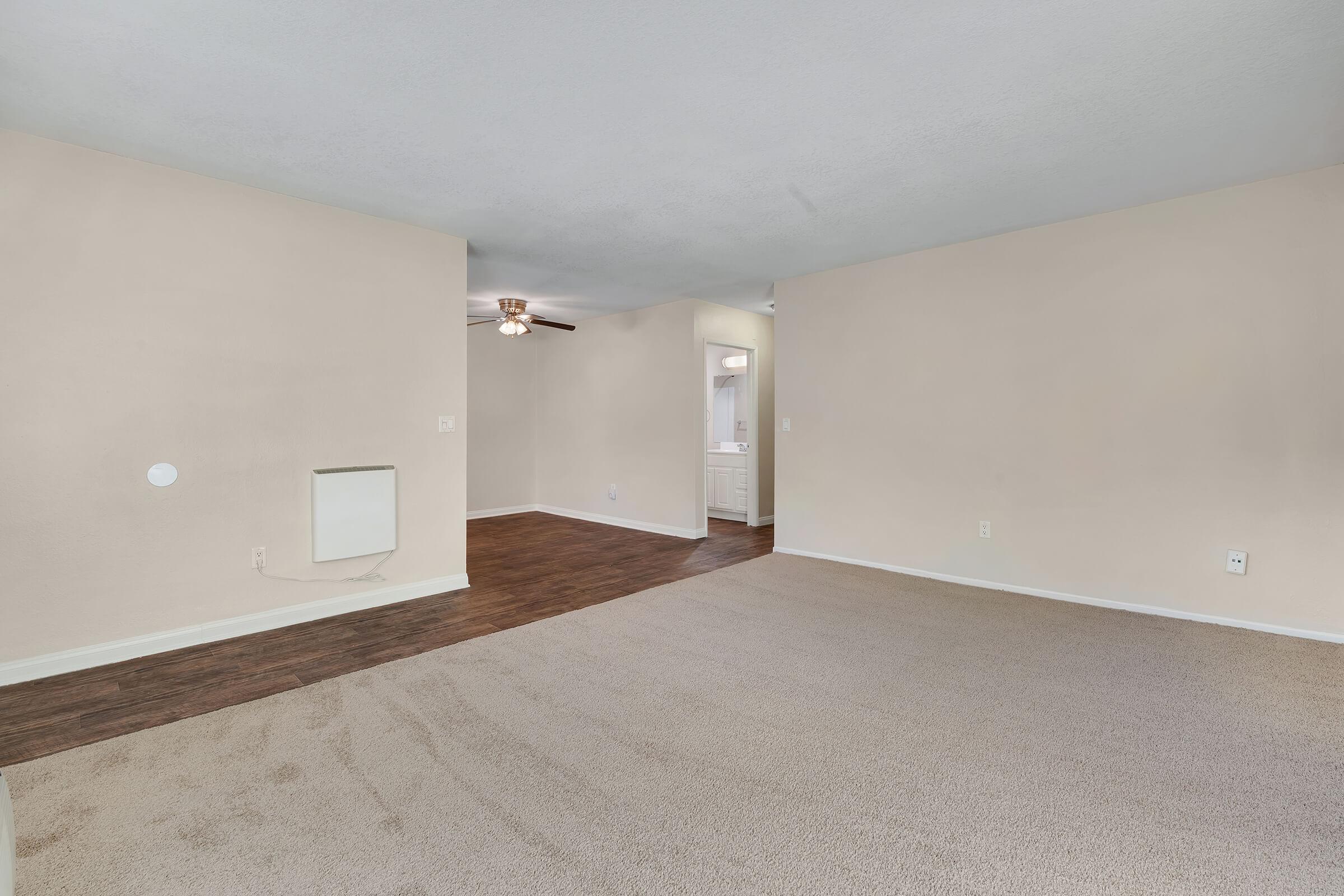 A spacious, empty living room featuring light beige walls and a ceiling fan. The floor is covered in light brown carpet, with a section of dark laminate flooring visible. A doorway leads to a hallway in the background, and there is a wall-mounted unit on the left side. The overall ambiance is bright and neutral.
