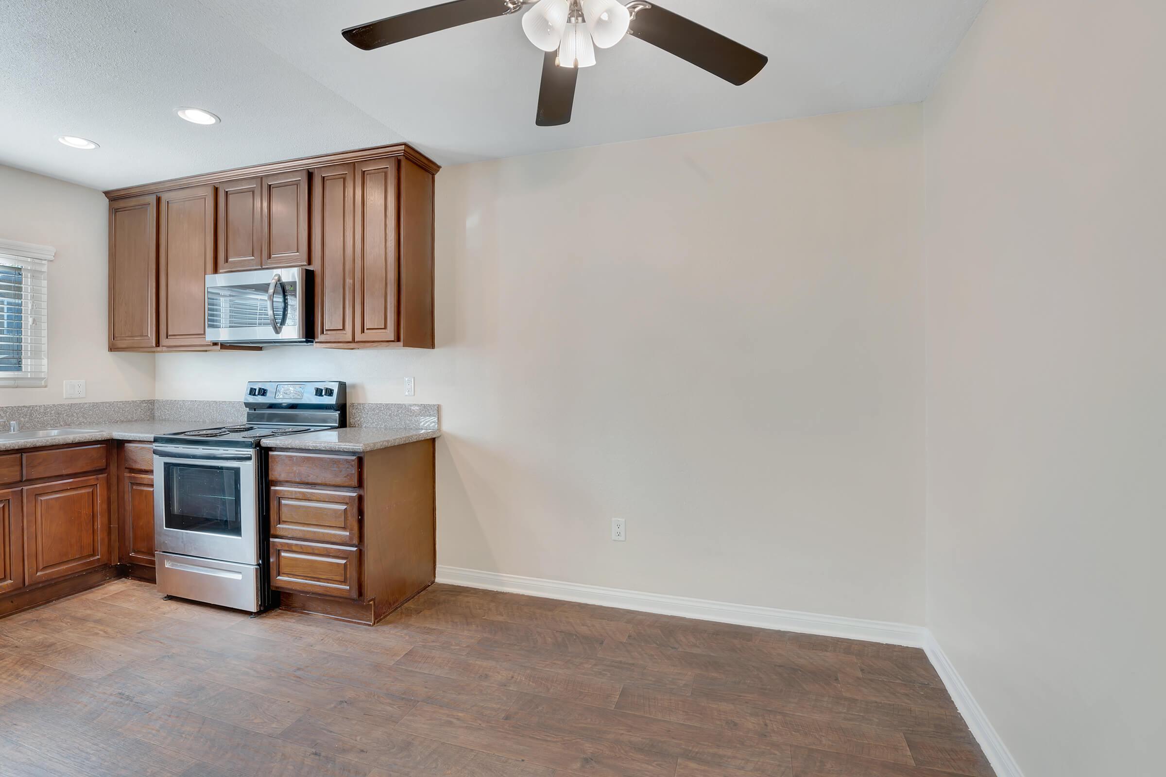 A clean, modern kitchen featuring dark wooden cabinets, a stainless steel oven, and a ceiling fan. The room has a light-colored wall and a spacious area, showcasing a well-lit, uncluttered space suitable for cooking and dining. The flooring is a warm wood finish.