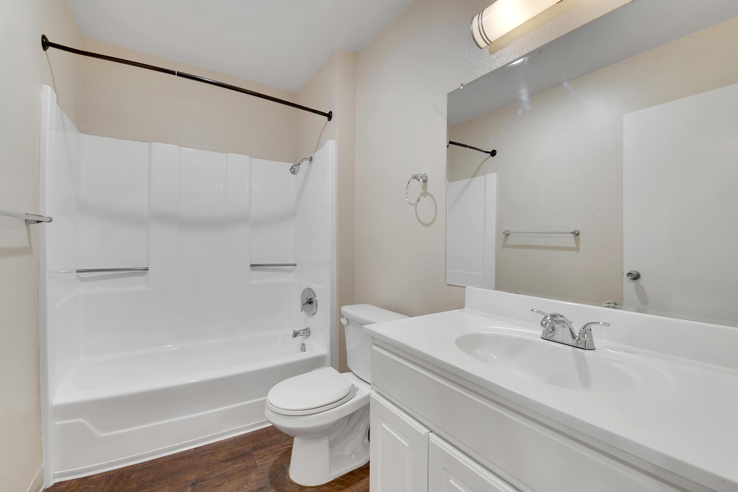 A clean, modern bathroom featuring a white bathtub with a shower curtain, a white toilet, and a sink with a countertop. The walls are neutral-colored, and there is a large mirror above the sink. The flooring appears to be a wooden texture, adding warmth to the space.