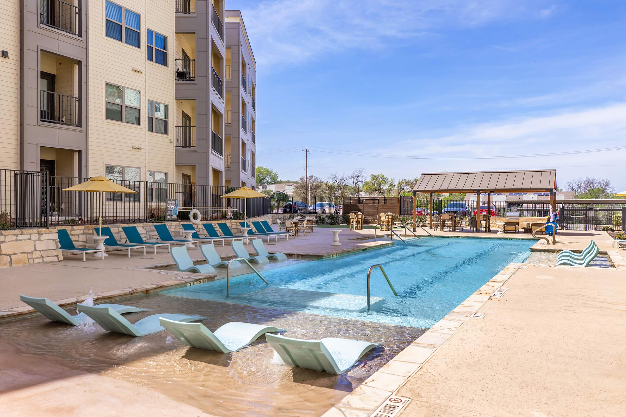 Swimming pool area featuring lounge chairs and umbrellas, with a clear blue sky above. The pool has a spacious deck with a few people enjoying the sun. There’s a BBQ area and outdoor furniture nearby, creating a relaxing atmosphere for recreational activities.