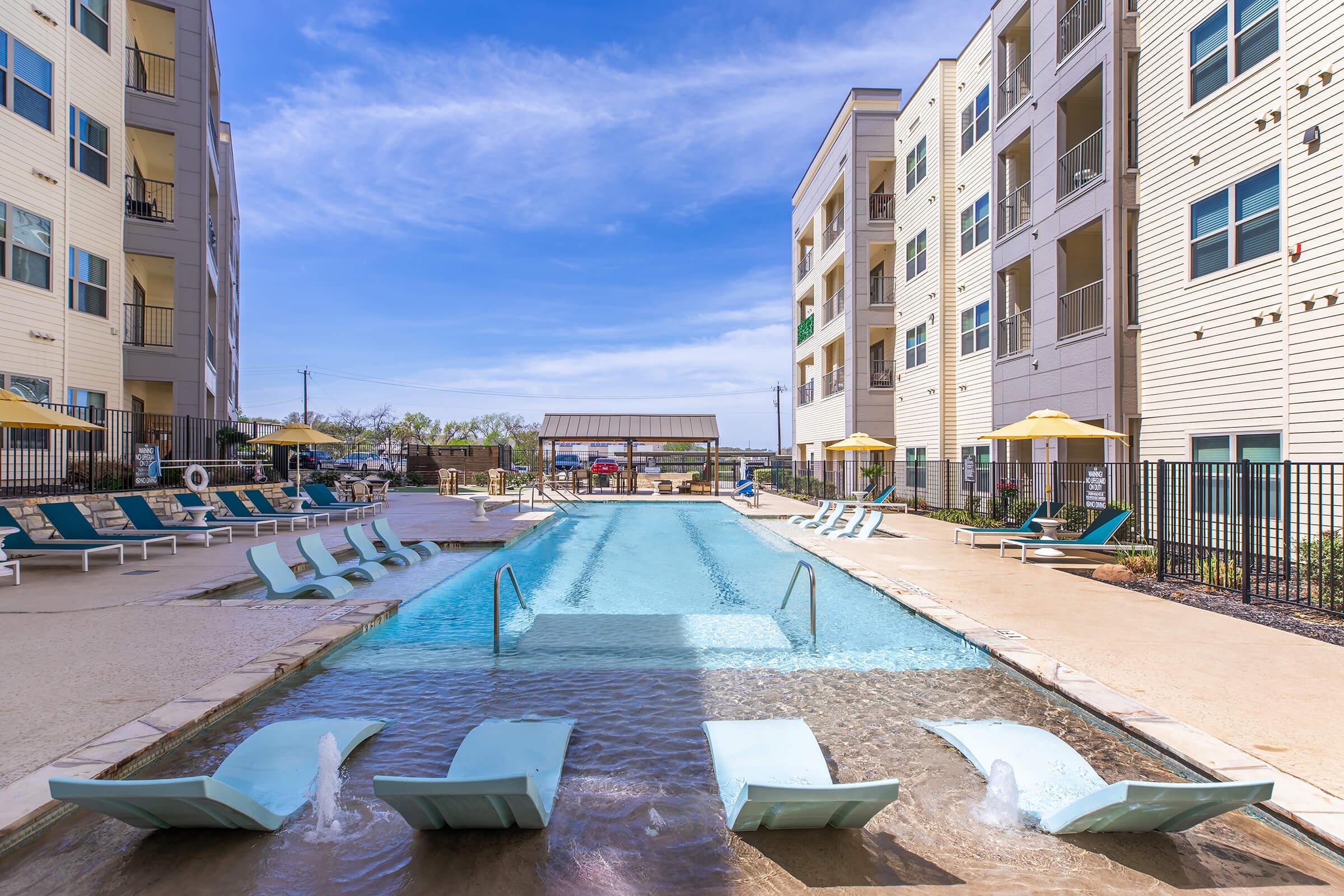 A modern apartment complex pool area featuring a clear blue swimming pool with poolside lounge chairs. Umbrellas provide shade, and there are nearby buildings with balconies visible. The sky is bright with a few clouds, creating a sunny atmosphere.
