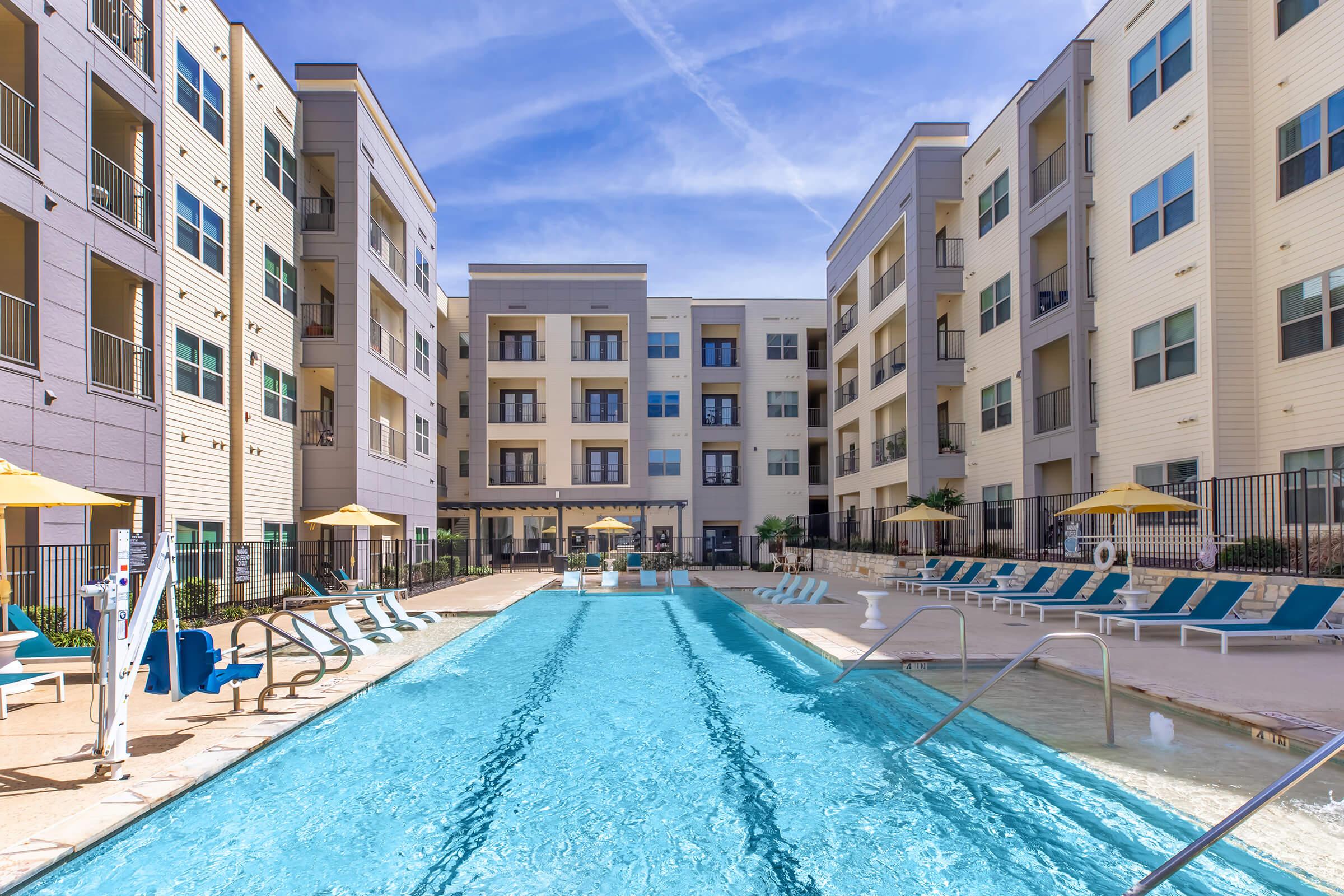 A modern apartment complex featuring a bright blue swimming pool surrounded by lounge chairs. The pool area is framed by multiple multi-story buildings with balconies. Umbrellas provide shade, and the sky is clear with a few clouds. The setting offers a relaxing outdoor atmosphere.