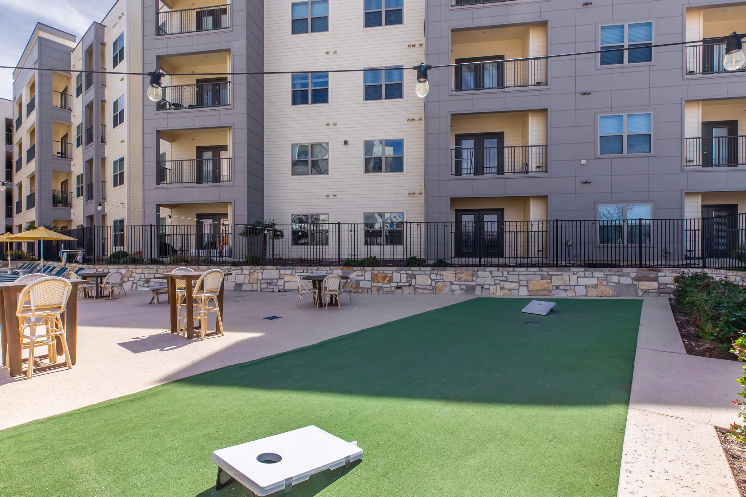 A well-lit outdoor area with a green lawn, featuring a cornhole setup, lounge chairs, and small tables. In the background, a multi-story apartment building with balconies is visible, surrounded by decorative landscaping and string lights overhead.