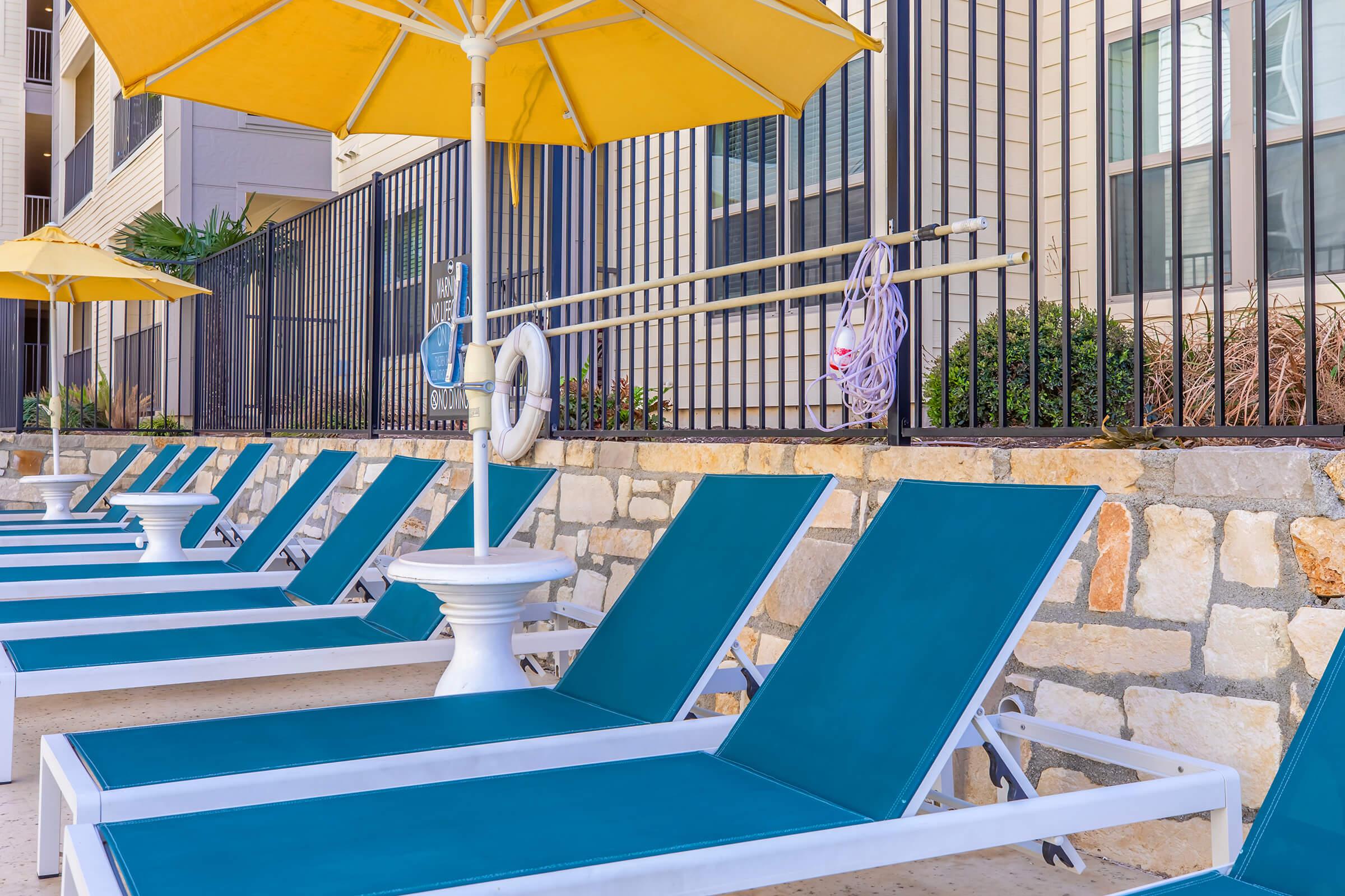 Lounge chairs with teal cushions lined up by a swimming pool, under yellow umbrellas. A white life preserver hangs nearby, and a fence runs along the background, with some greenery visible. The setting appears to be a residential or resort pool area.