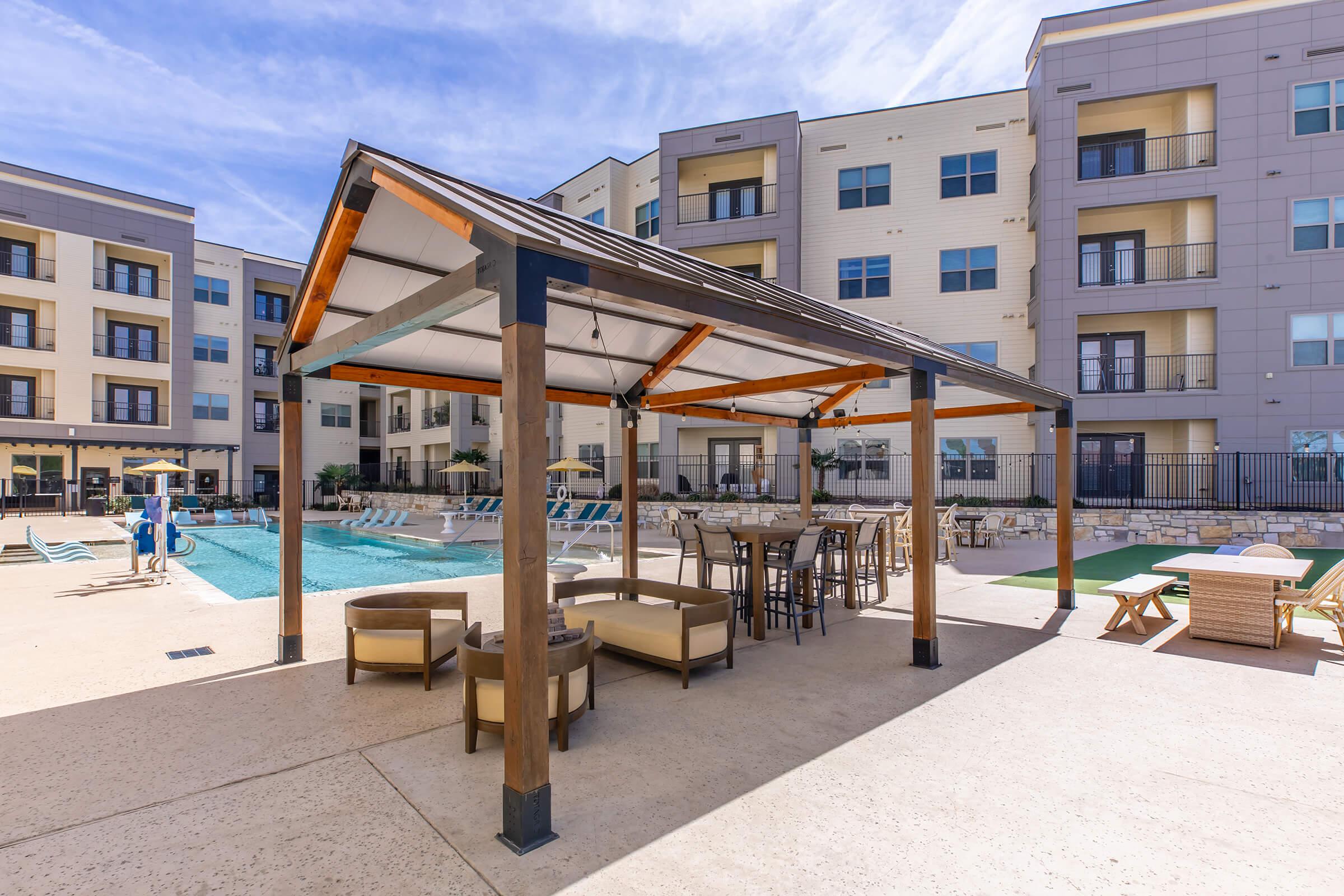 Outdoor pool area featuring a spacious lounge with seating under a covered pergola, surrounded by a modern apartment building. Sun loungers and umbrellas line the poolside, creating a welcoming atmosphere for relaxation and socializing. Clear blue sky enhances the resort-style ambiance.